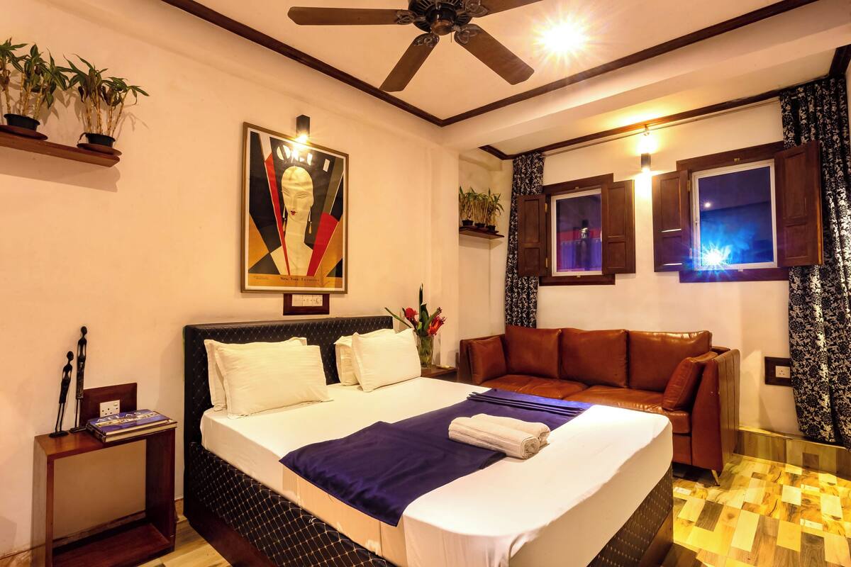 A hotel room with a bed, a brown leather sofa, a ceiling fan, a painting on the wall, and a window with curtains.