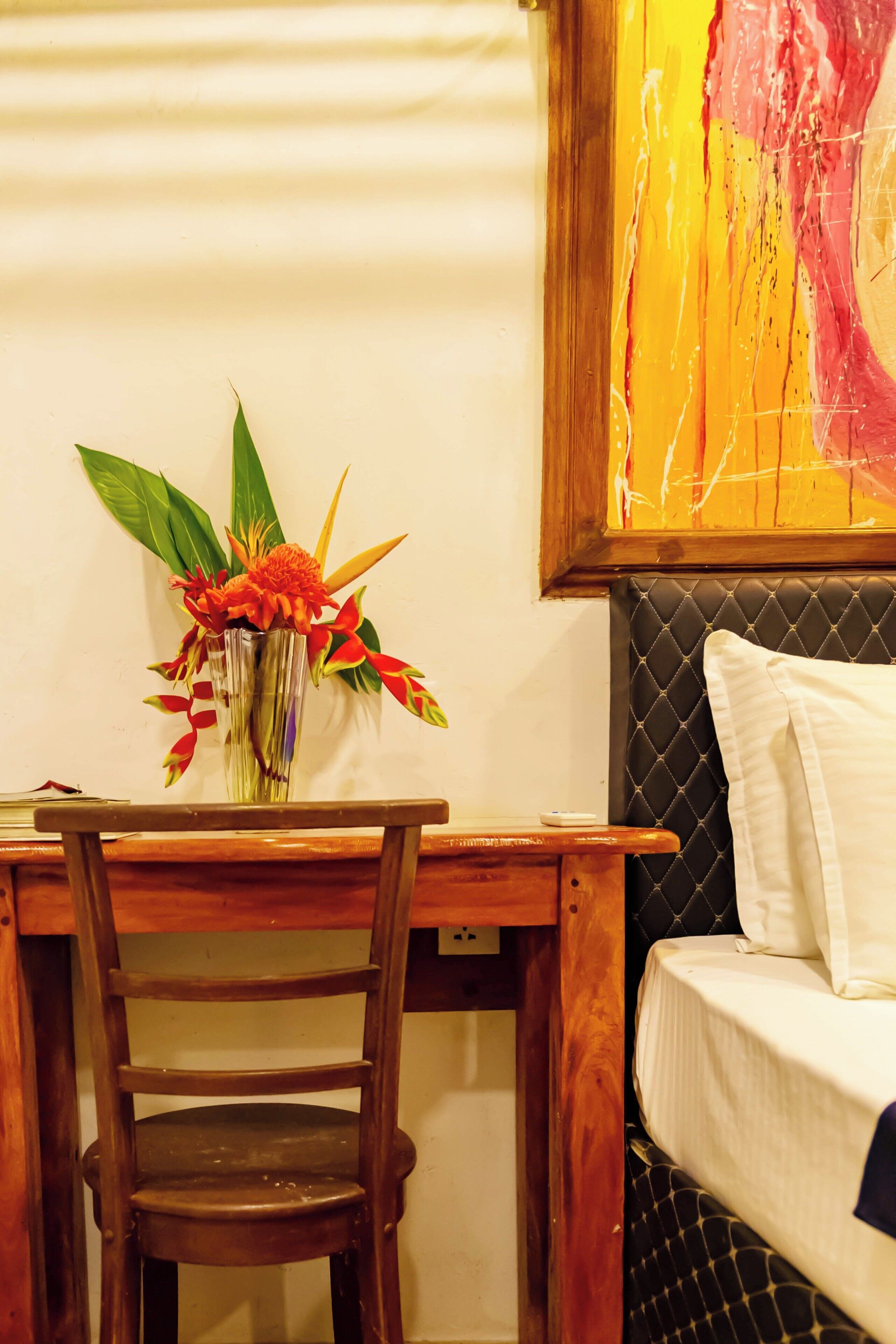 A wooden desk with a vase of flowers, a chair, and a bed with a quilted headboard.