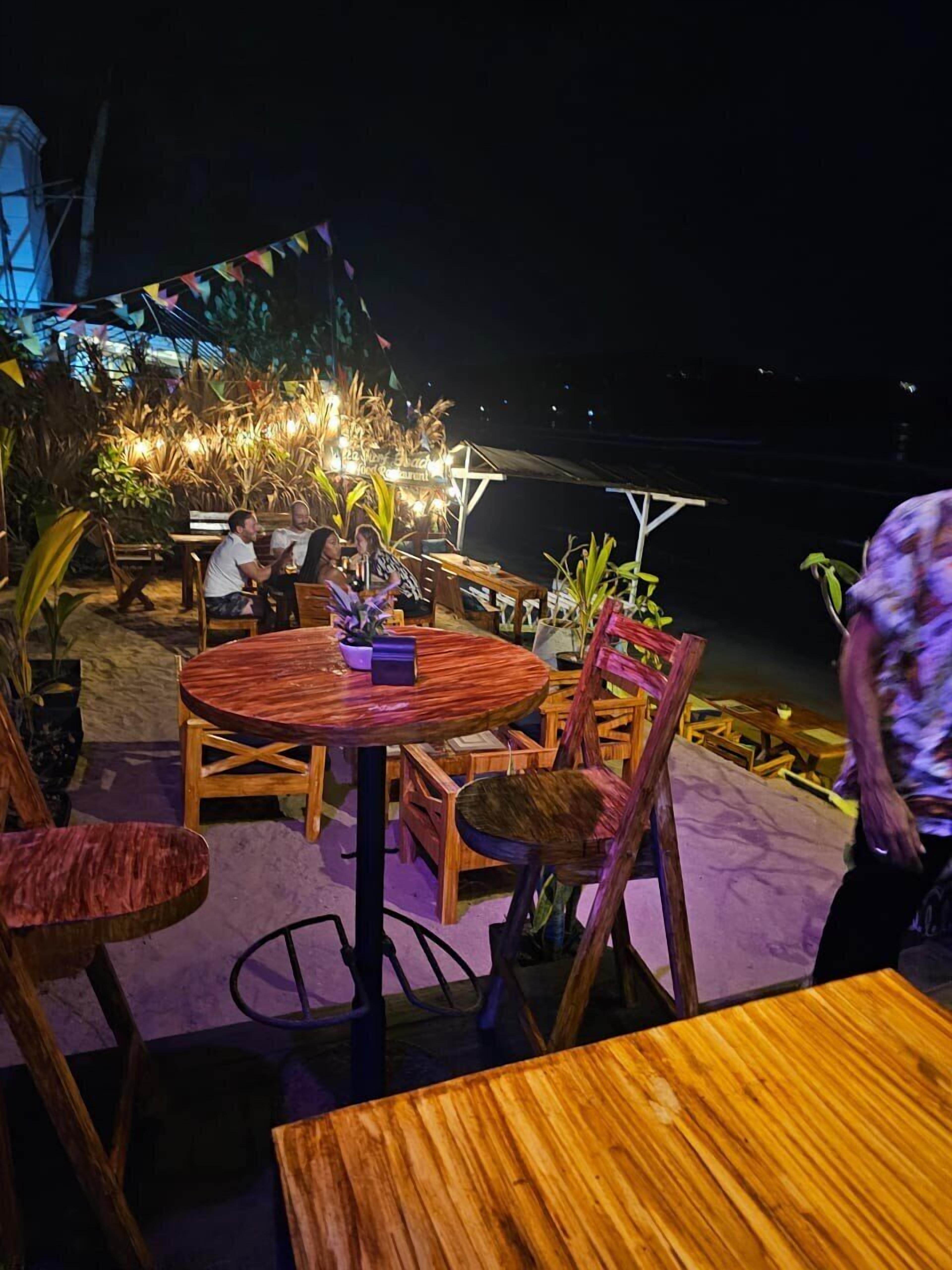 Un espacio de comedor al aire libre con mesas y sillas de madera, rodeado de plantas y luces colgantes.