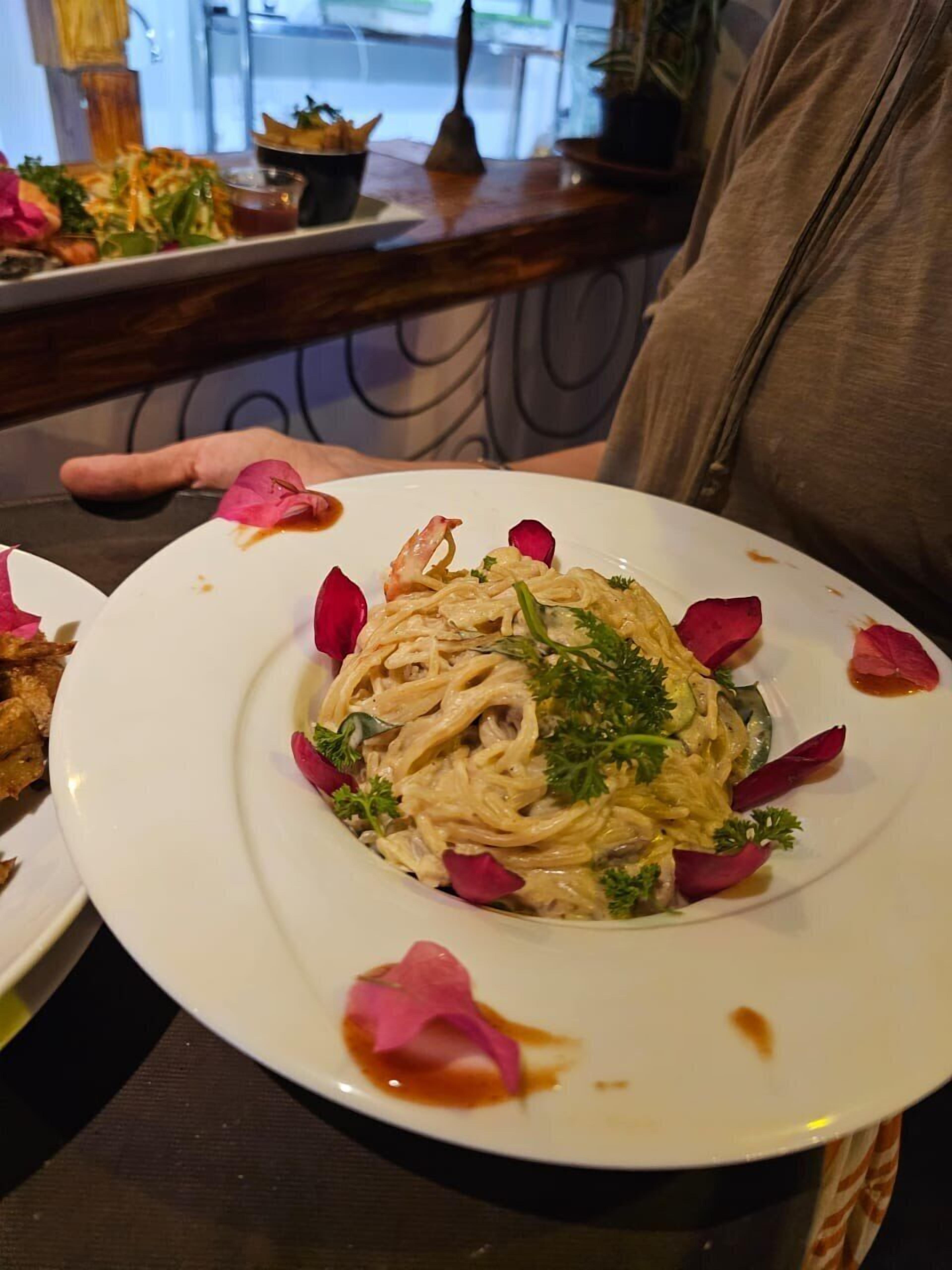 Un plato de pasta con salsa cremosa, decorado con perejil y pétalos rojos, sobre un plato blanco.
