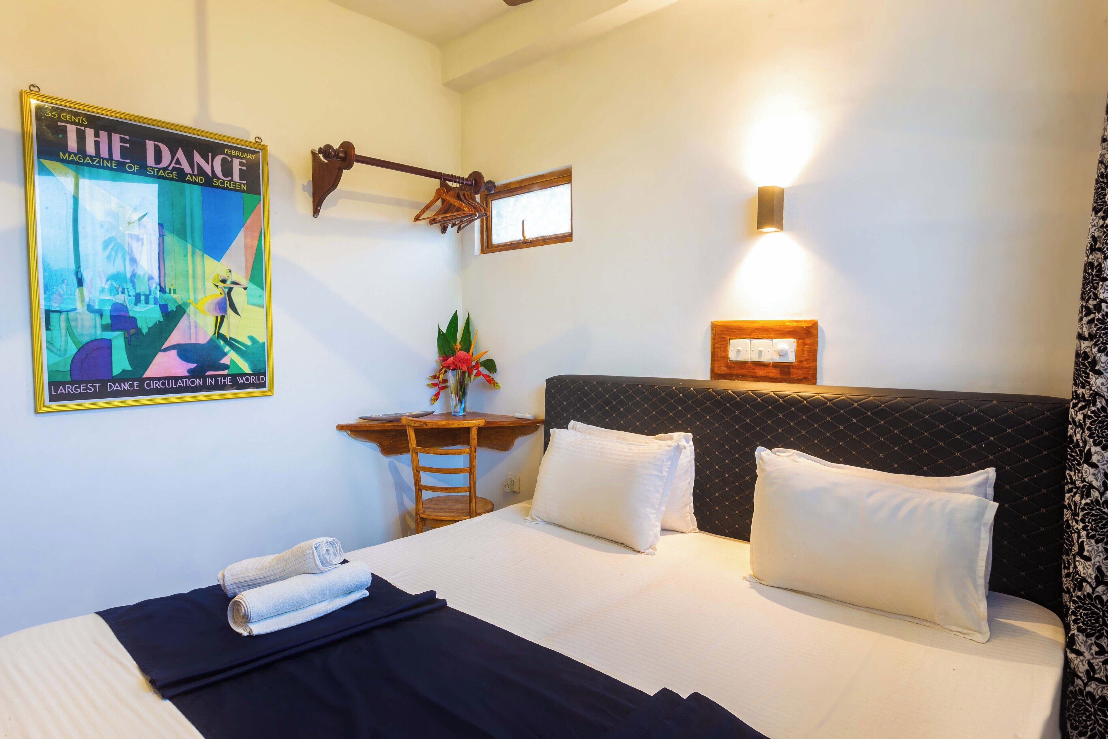 A hotel room with a bed, a desk, a chair, a wall-mounted light, and a poster.