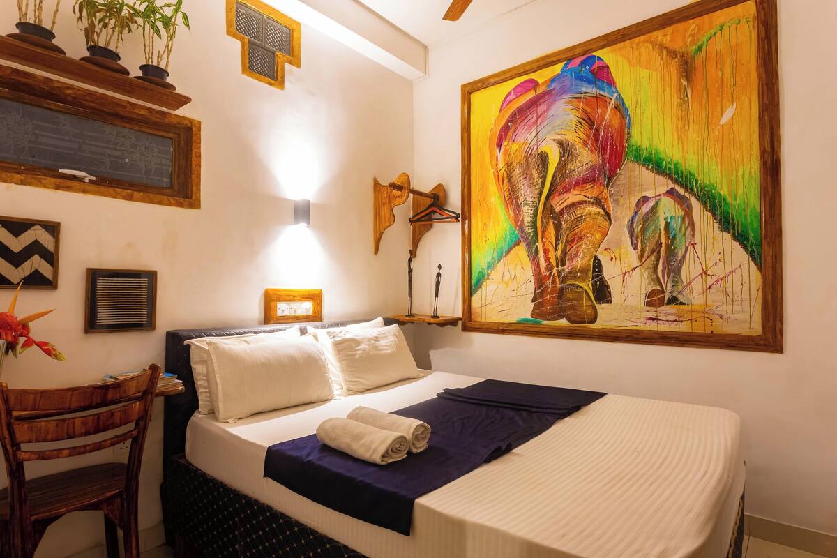 A hotel room with a bed, a desk, a chair, a wall-mounted shelf with potted plants, a wall-mounted speaker, and a large colorful painting.