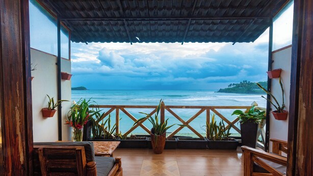 A wooden deck with a view of the ocean, potted plants, and a wooden table and chair. - Villa Surf Beach (Galle)