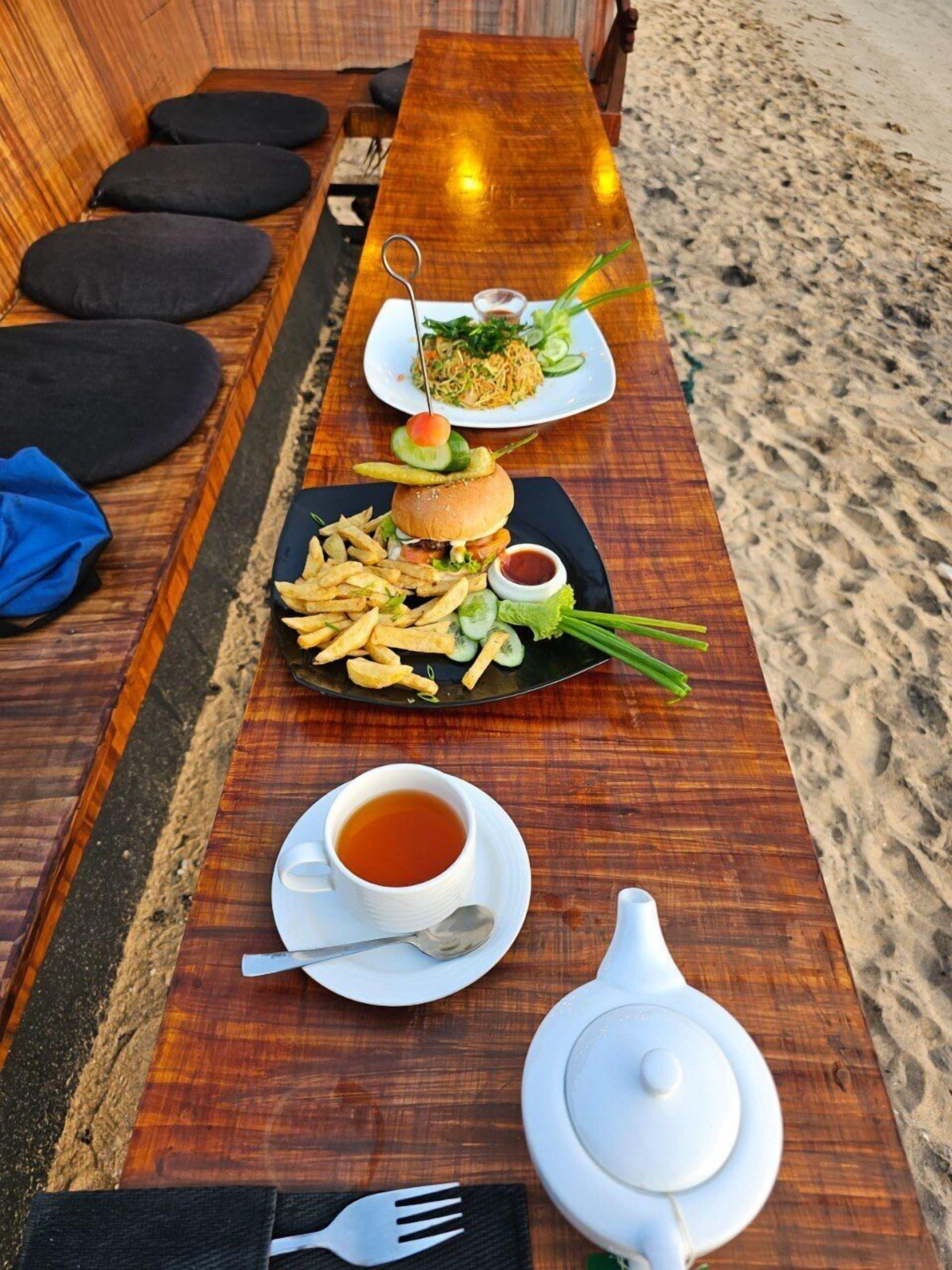 A wooden dining table with a meal consisting of a burger, fries, and a side salad, along with a cup of tea and a teapot.