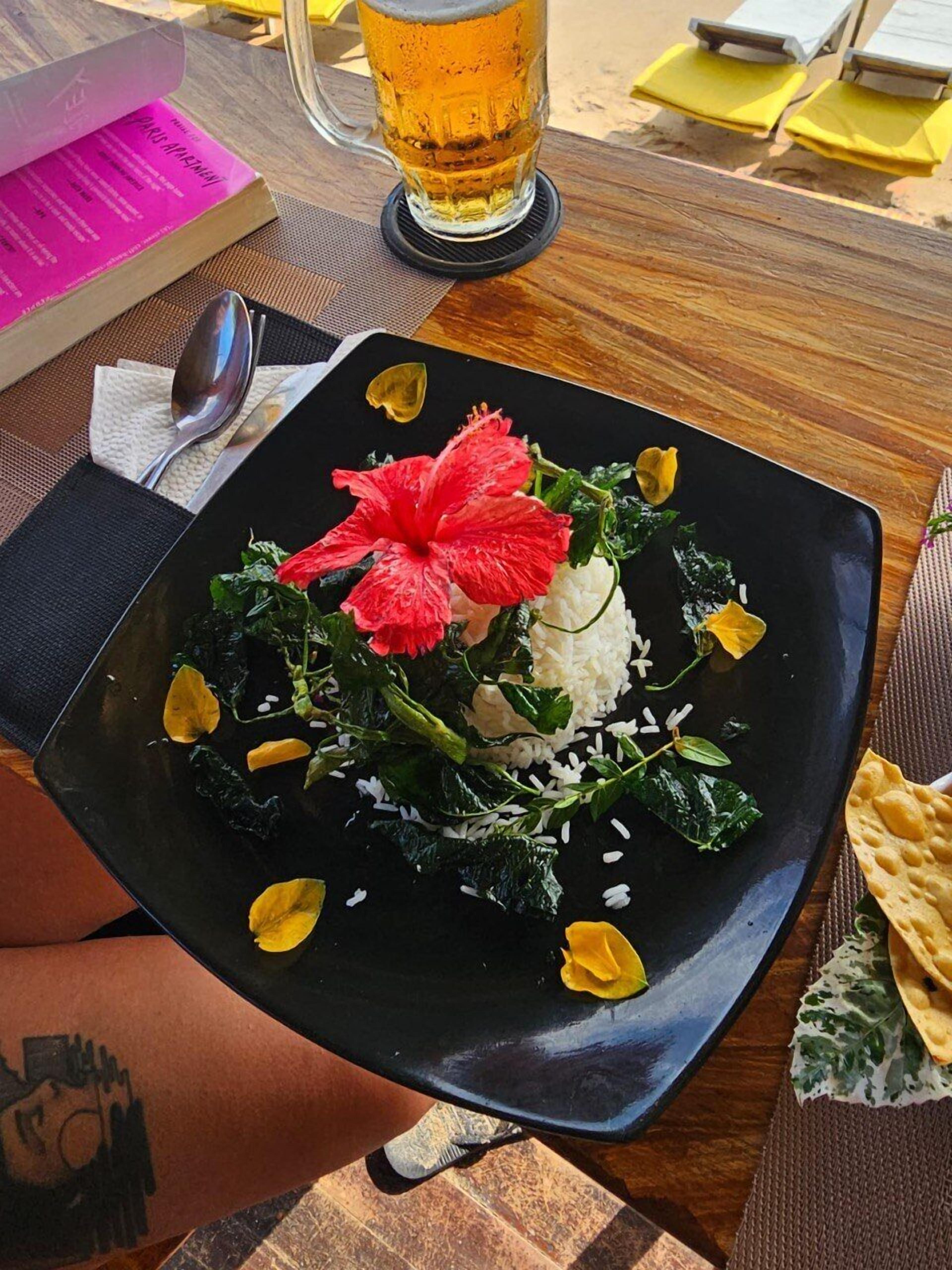 A black plate with a salad, a red flower, and rice, held by a person with a tattoo.