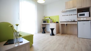 Een moderne keuken met houten kastjes, een magnetron en een koelkast. In de woonkamer staat een eettafel met stoelen en een groene bank.