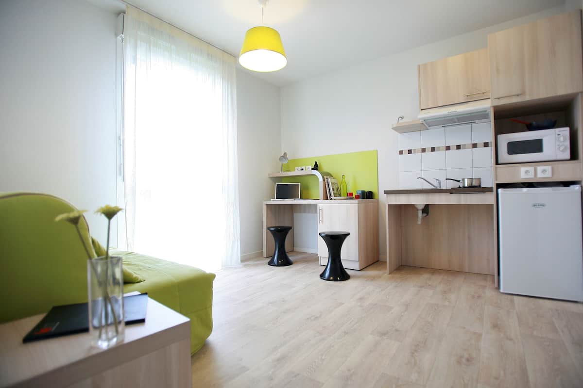 Une cuisine moderne avec des meubles en bois, un four à micro-ondes et un réfrigérateur. On y trouve une table à manger avec des chaises, ainsi qu’un canapé vert dans l’espace salon.