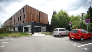 Een modern gebouw met een plat dak en grote ramen, een parkeerplaats met aangegeven parkeerplaatsen en een bord met daarop 'verboden te parkeren'.