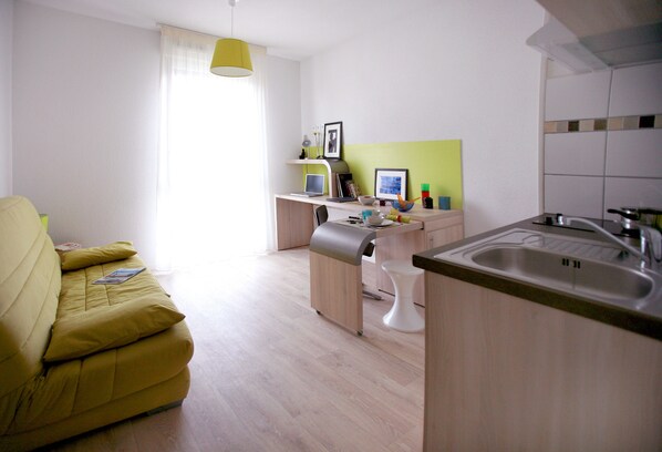A modern, well-lit living room with a yellow sofa, a small dining area, and a kitchen with a sink and countertop.