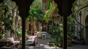 Un patio exterior con zona de estar, que incluye mobiliario de mimbre y una mesa, rodeado de abundante vegetación y arcos arquitectónicos.