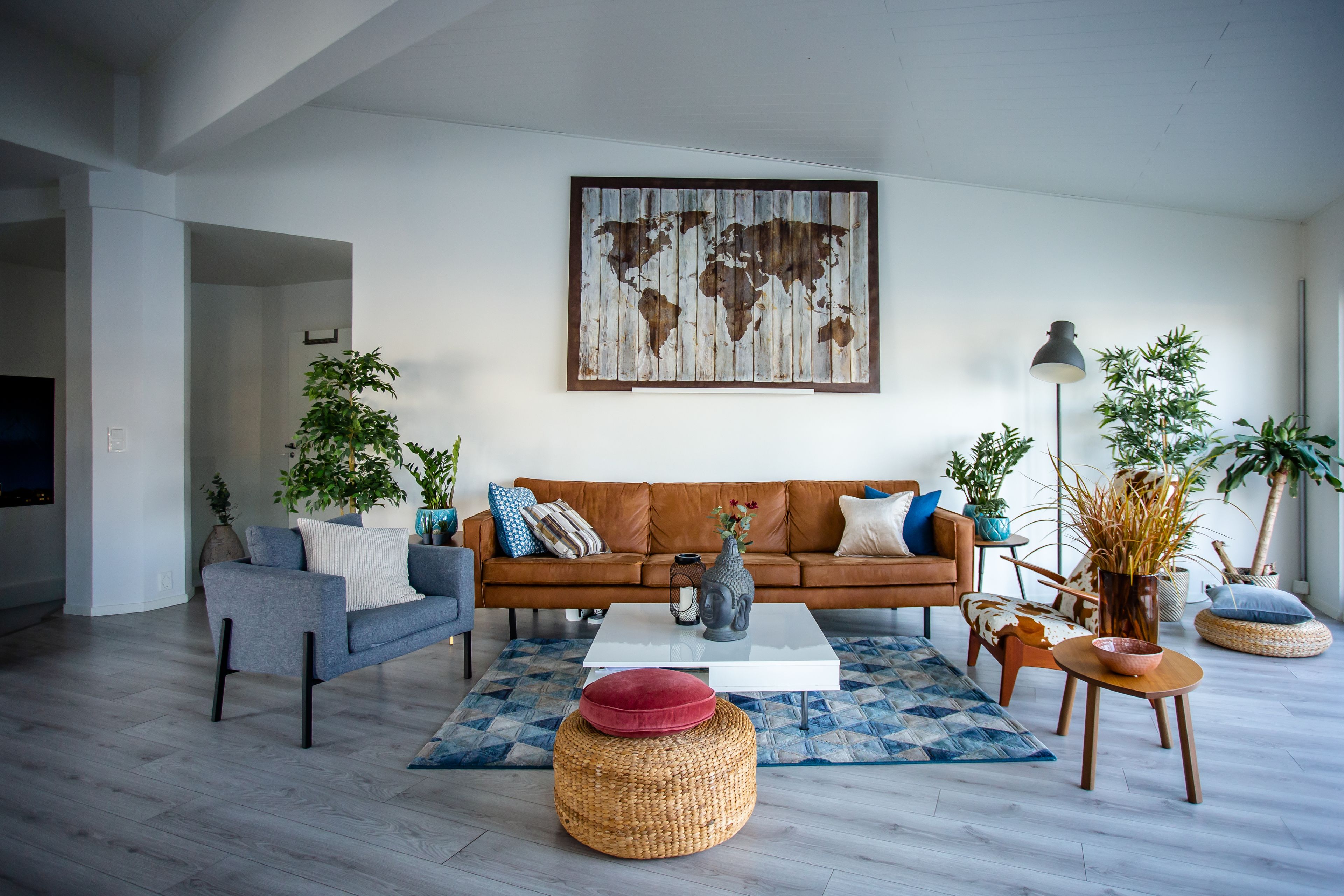 A modern living room with a brown leather sofa, a grey armchair, and a coffee table. There are two potted plants, a world map artwork on the wall, and a floor lamp.