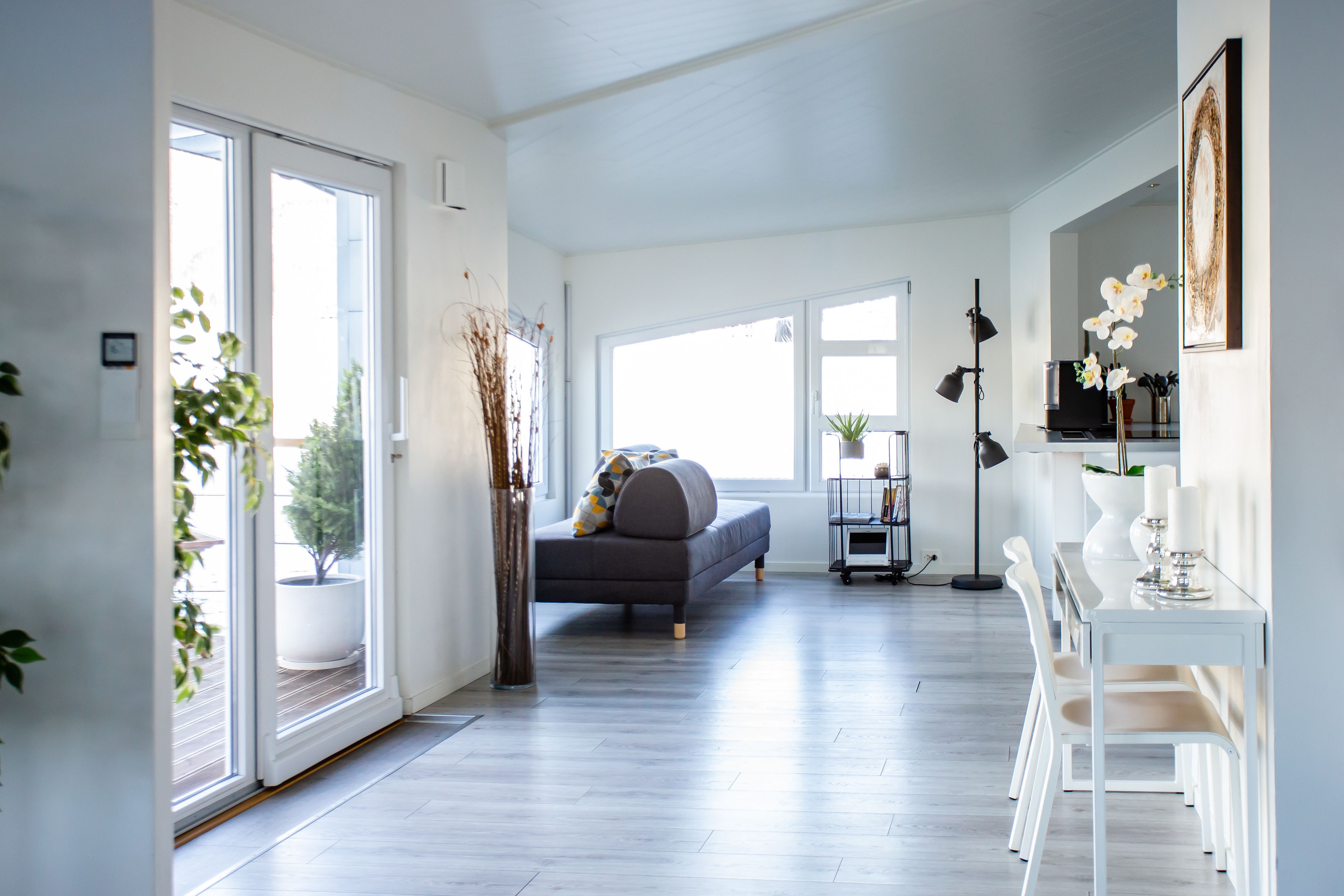 A modern living room with a grey sofa, white chairs, a dining table with candles, and large windows.