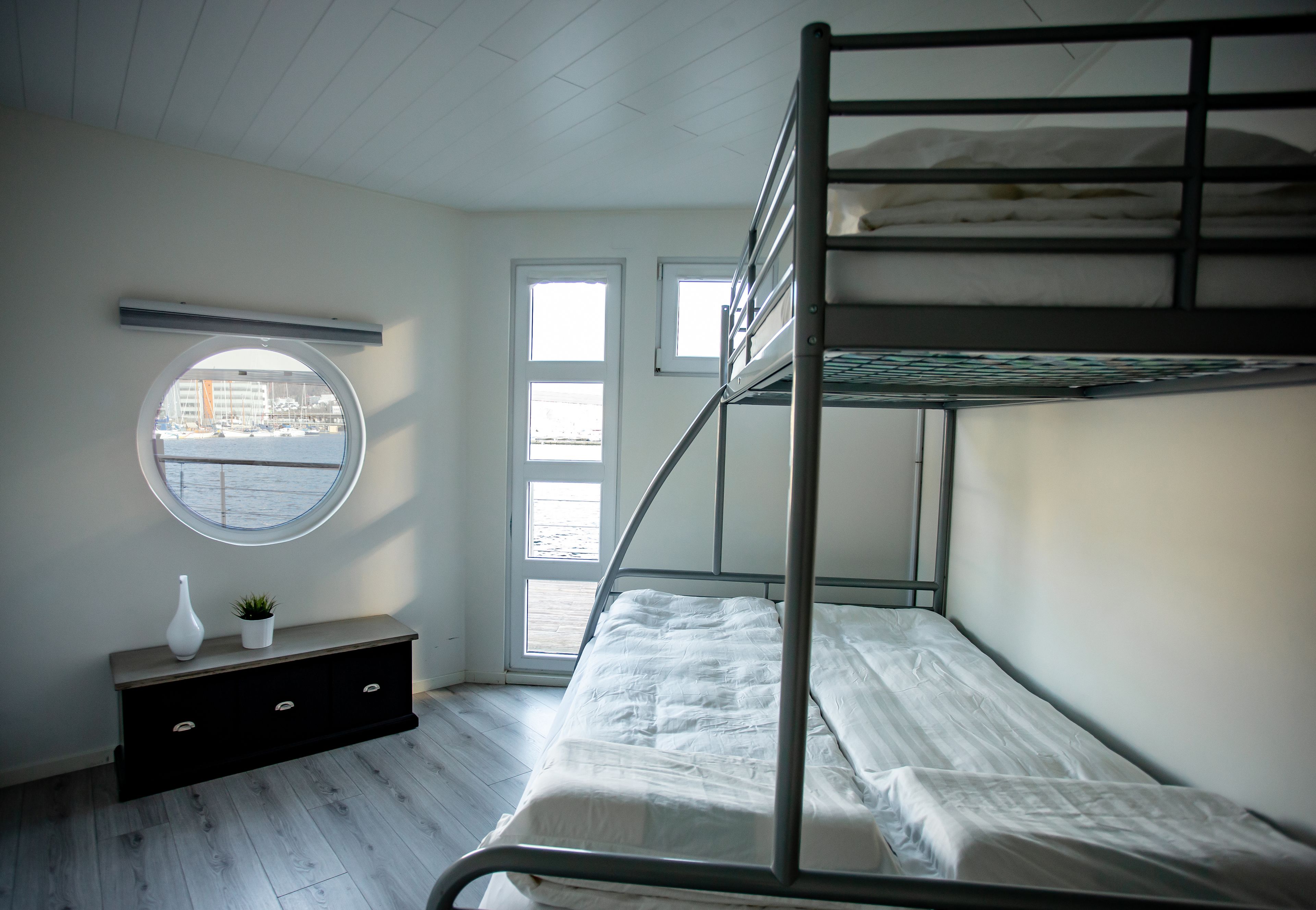 A bunk bed room with a round window, a small plant, and a white vase on a dark wooden dresser.