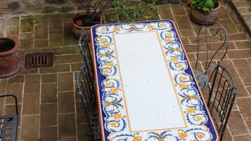 A rectangular outdoor table with a decorative border, surrounded by metal chairs, set on a stone patio with potted plants and a metal fence.