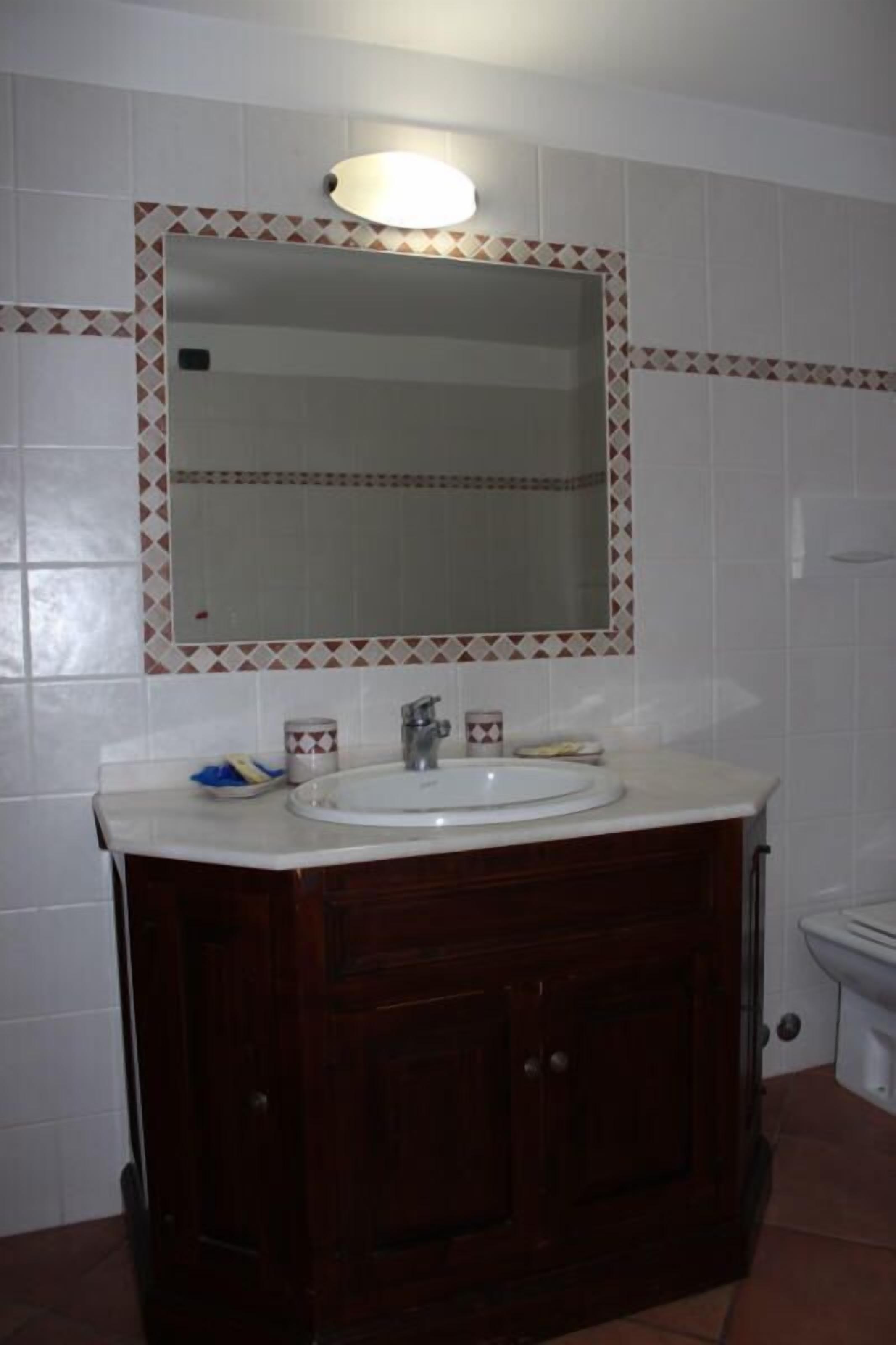 Een badkamer met een witte wastafel, een donker houten wastafelmeubel en een spiegel met een geometrisch tegelreliëf.