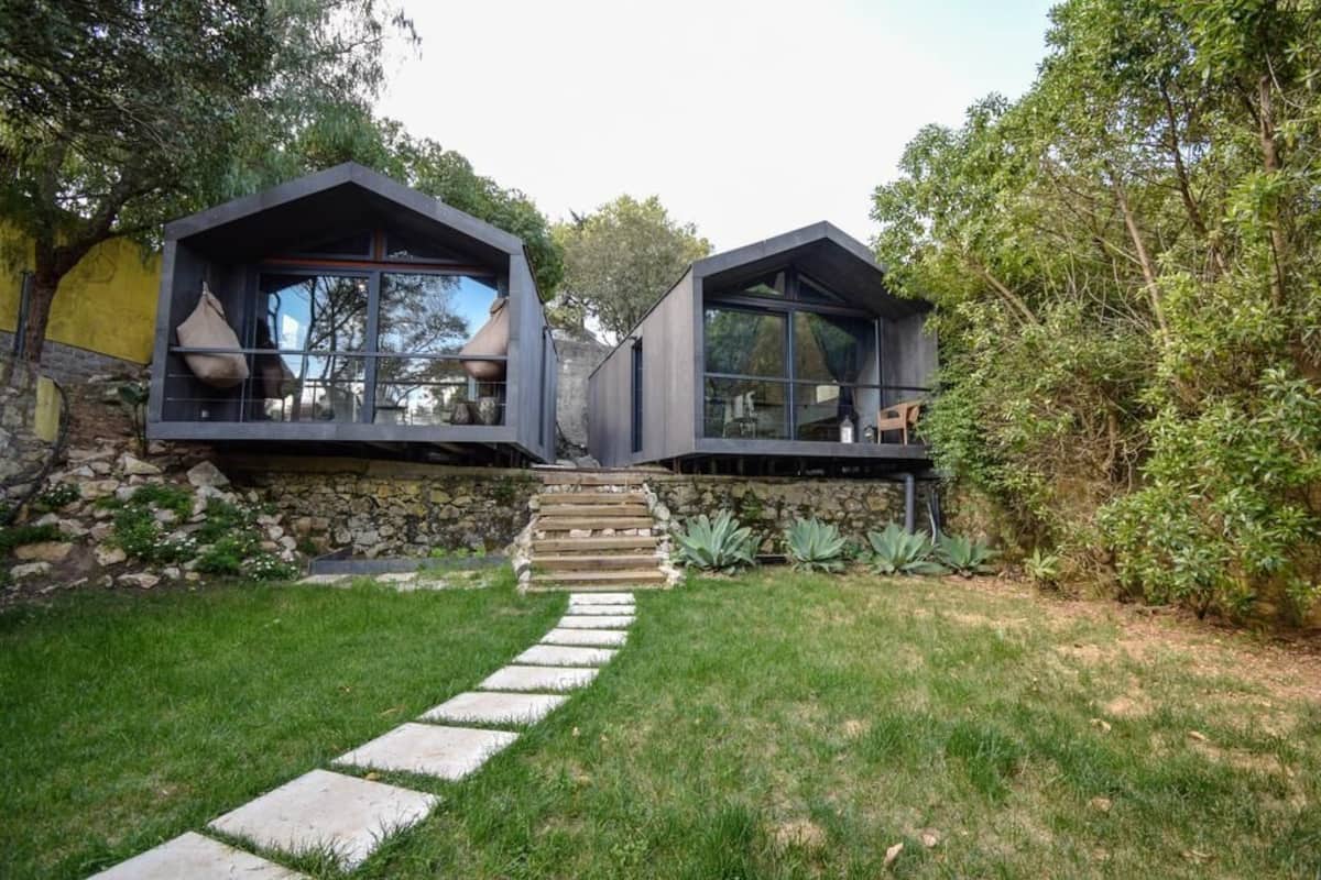 A modern house with two large, dark-colored rooms with large windows, a stone wall, and a stone staircase leading to the entrance.