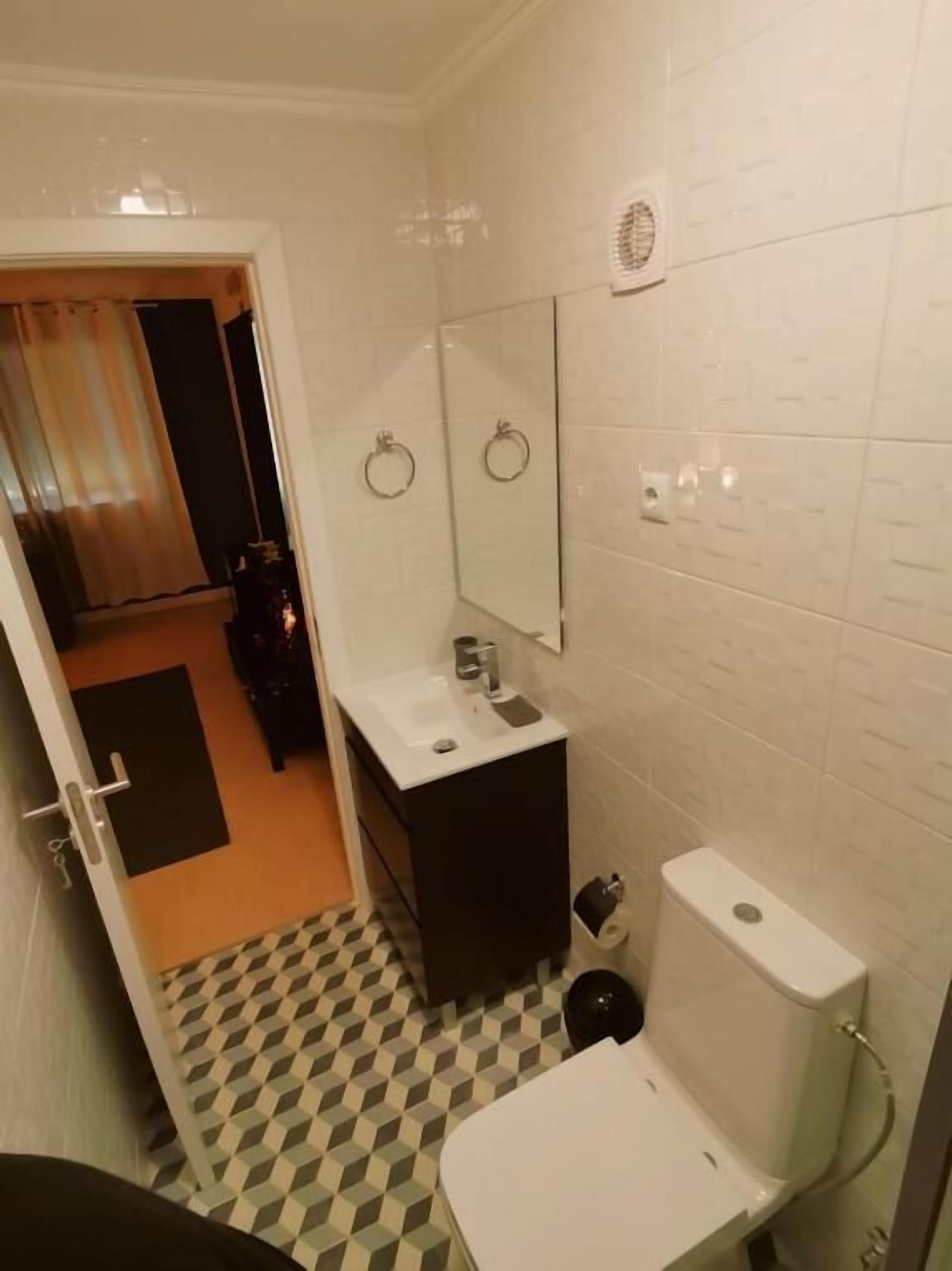 Un bagno piccolo con un lavandino bianco, uno specchio e un water. Il pavimento presenta un motivo a scacchi e una porta che dà su un'altra stanza.