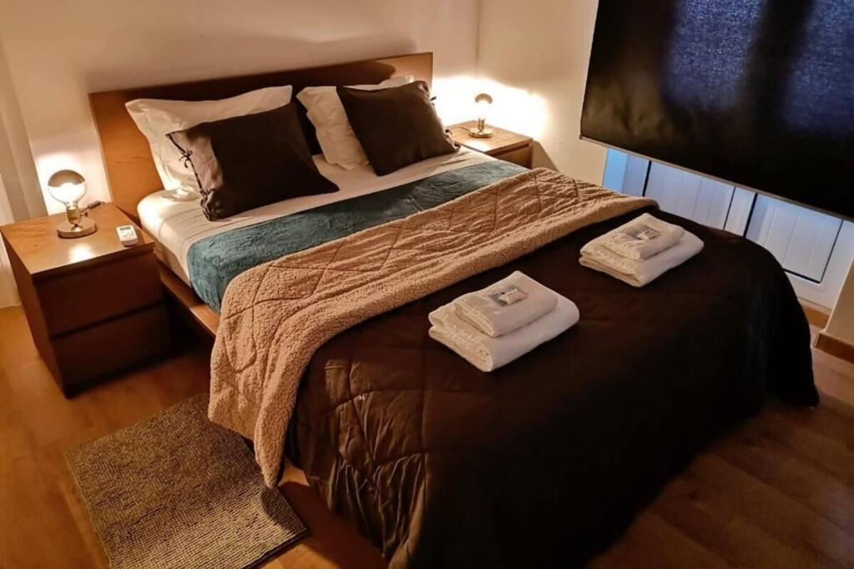 A neatly made bed with a brown cover, two pillows, and a folded towel on the bed. There is a nightstand with a lamp and a small table beside the bed.