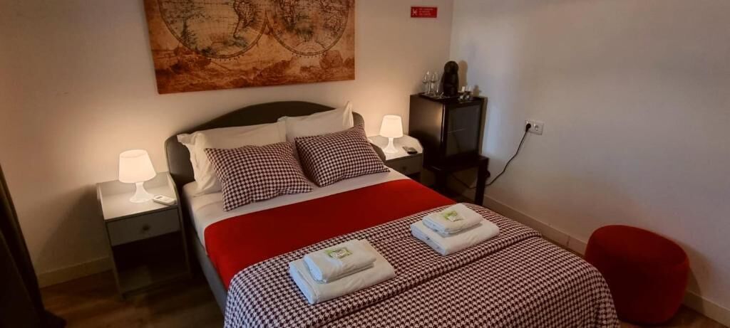 Een net opgemaakt bed met geruit beddengoed, twee bedlampen, een rode poef en een kaart aan de muur.