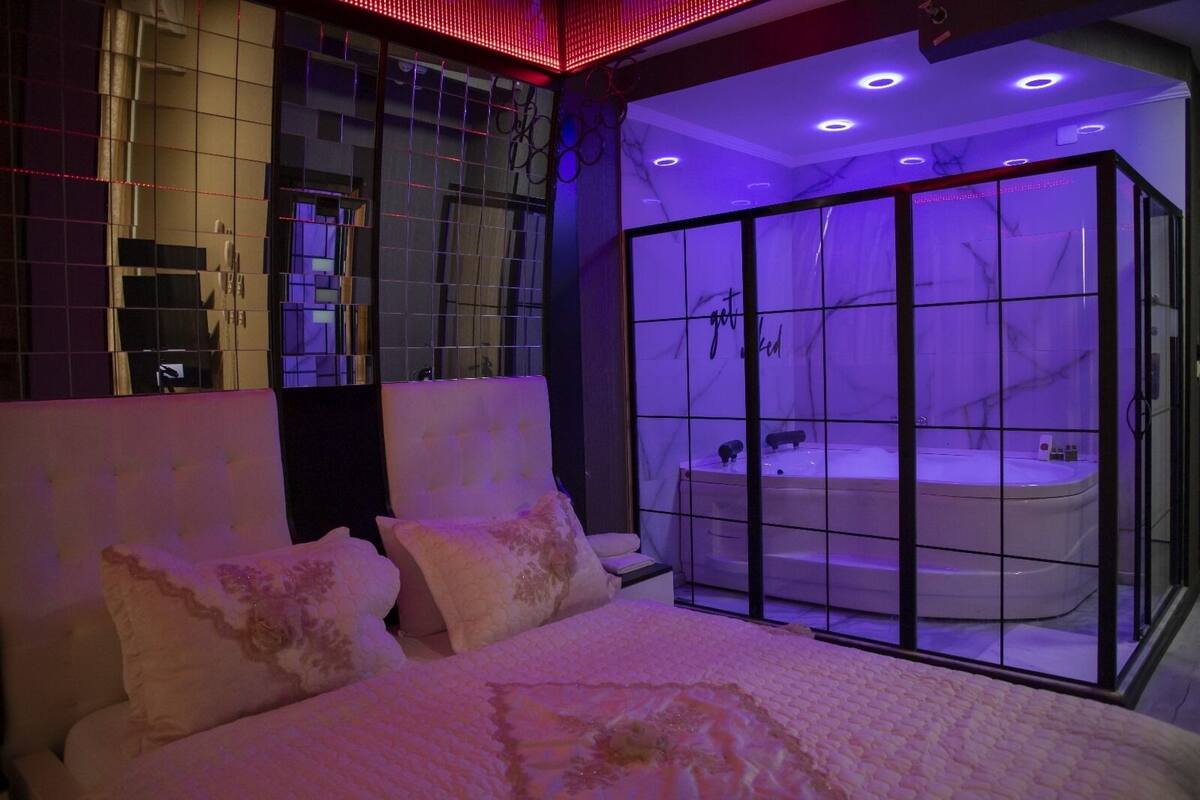 A modern bedroom with a large bed, a glass-enclosed jacuzzi, and a bathroom with a vanity and mirror.