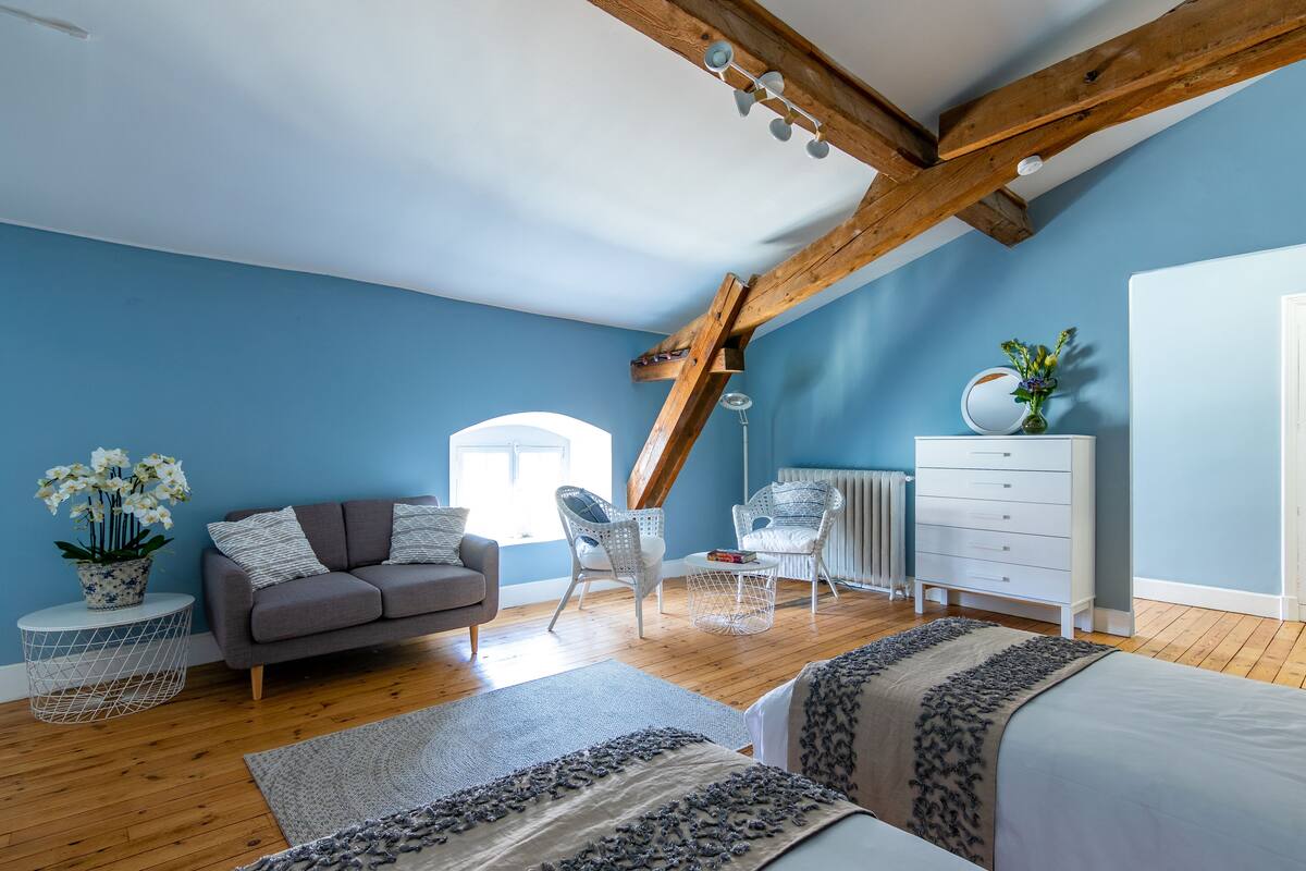 A room with a sloped ceiling, a grey sofa, white chairs, a white dresser, and a large window.