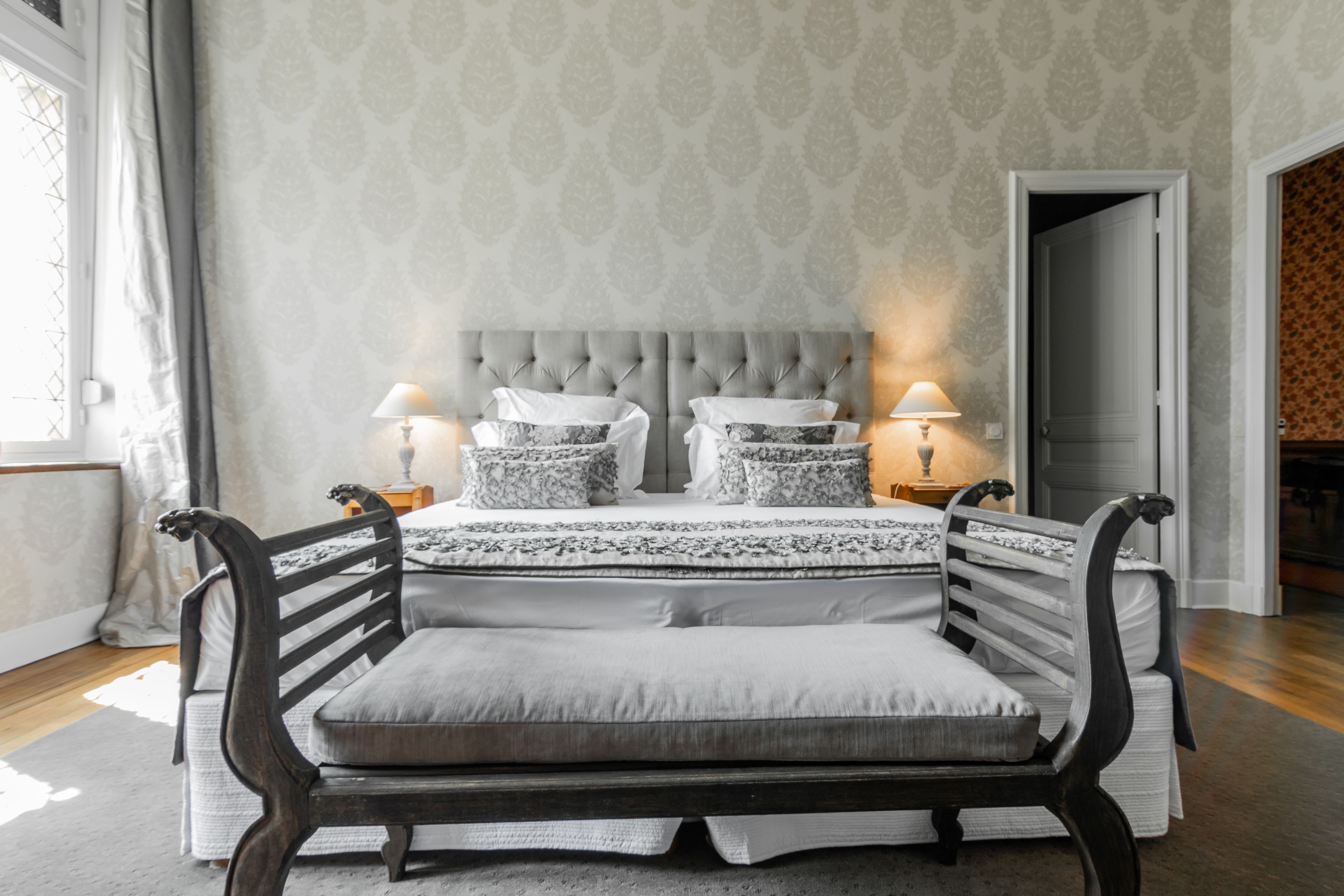 A bedroom with a tufted headboard, two bedside tables with lamps, a large bed with a patterned bedspread, and a dark wooden bench.
