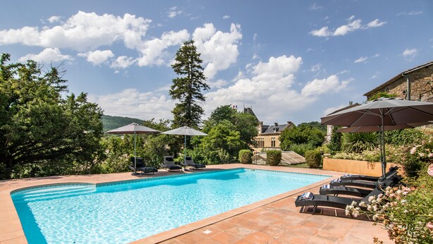 Ein rechteckiger Swimmingpool mit klarem, blauem Wasser, umgeben von einer gefliesten Terrasse. Liegestühle und Sonnenschirme bieten Schatten.