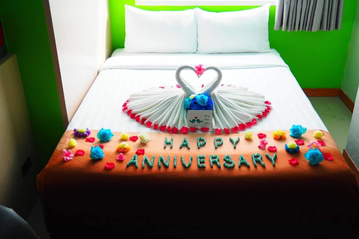 A bed with a heart-shaped decoration made of white feathers and a blue flower, surrounded by colorful flowers and the text "HAPPY ANNIVERSARY".