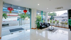 A modern hotel lobby with a reception desk, seating area, and large windows offering a view of the street and greenery outside. - Blue Orchids Hotel (Hat Yai)