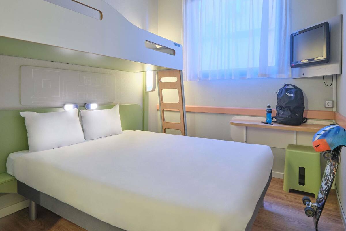 Ein Hotelzimmer mit einem Bett, einem Schreibtisch, einem Sessel, einem Fernseher, einem Fenster mit Vorhängen und einem Rucksack auf dem Schreibtisch.