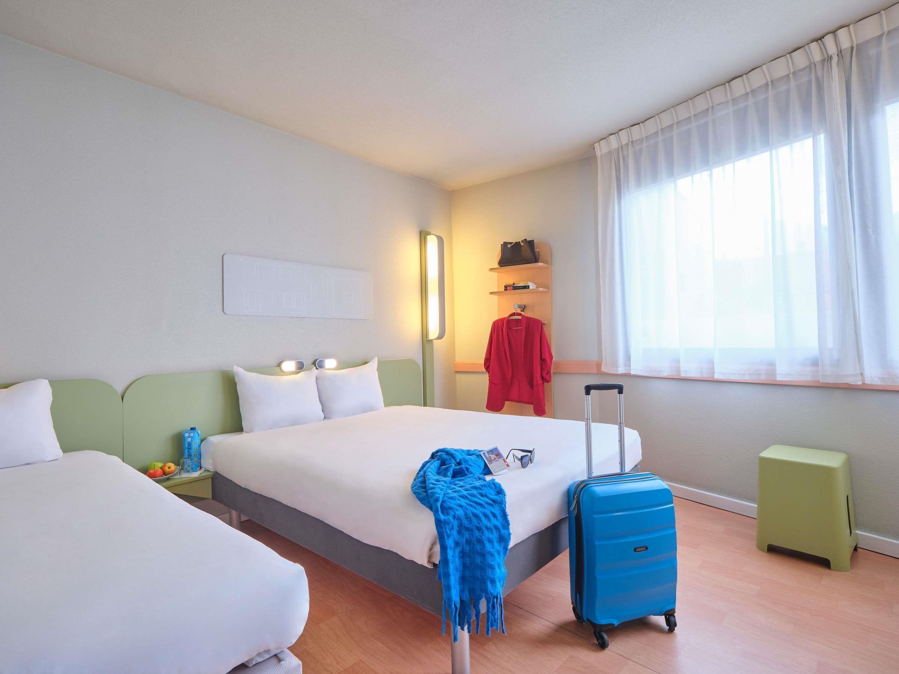 A hotel room with two beds, a nightstand, a suitcase, a small stool, a window with sheer curtains, and a wall-mounted shelf with a handbag.