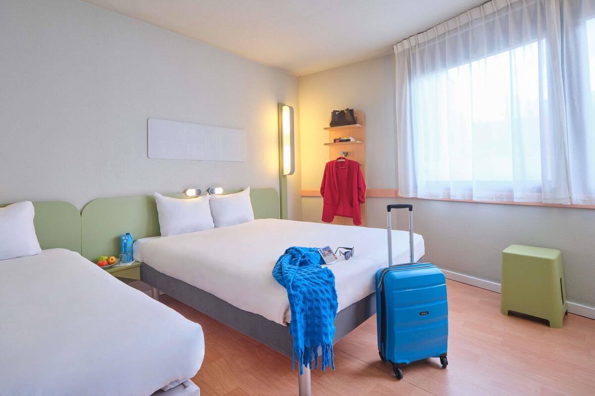 A hotel room with two beds, a nightstand, a suitcase, a small stool, a window with sheer curtains, and a wall-mounted shelf with a handbag.