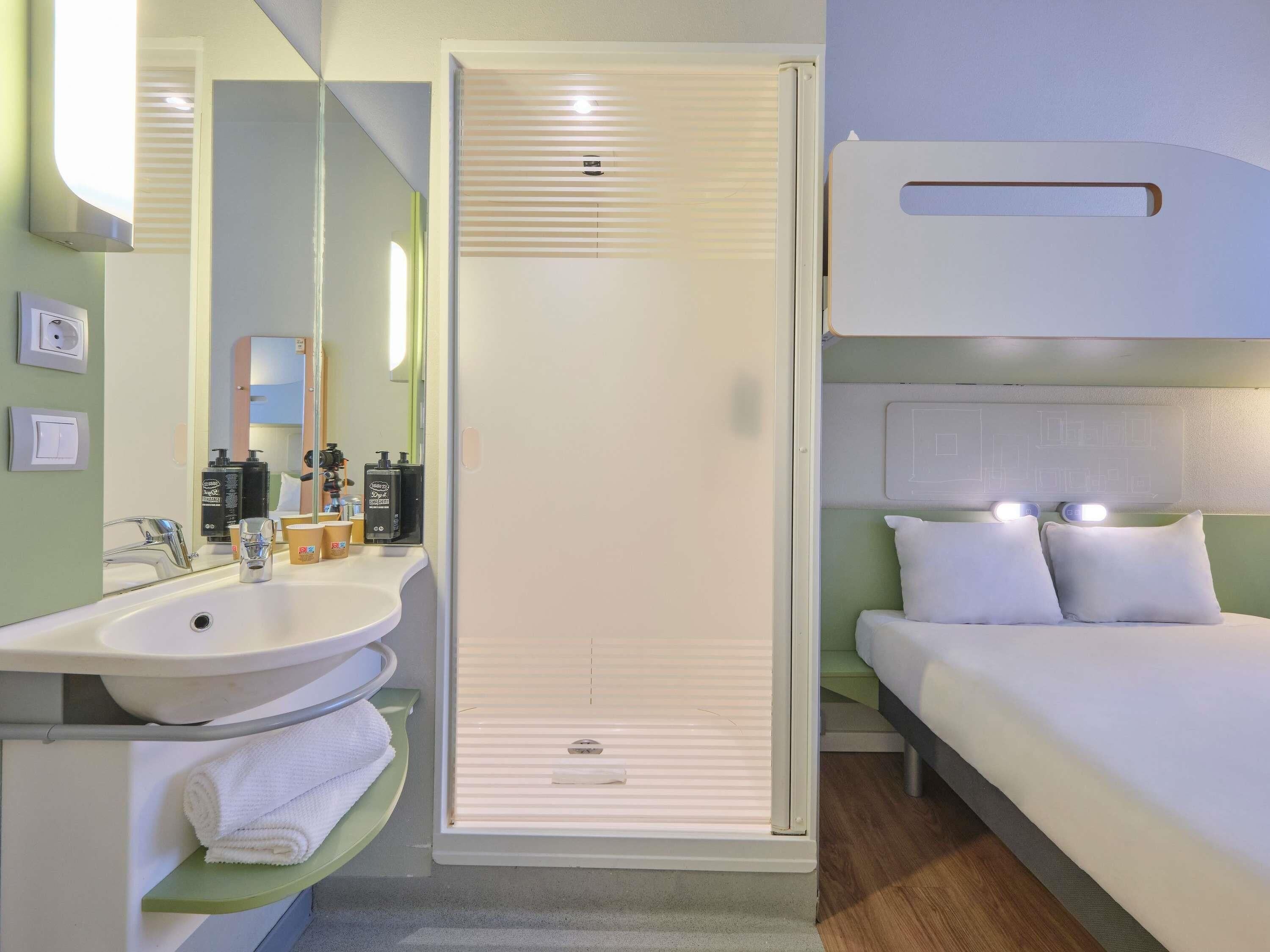 A hotel room with a white bed, a white sink, a mirror, and a shower with blinds.