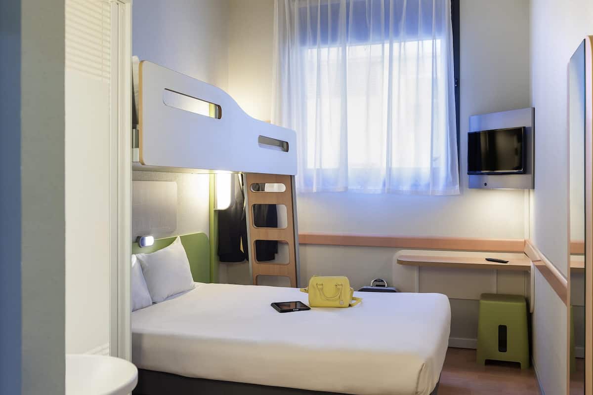 A compact hotel room with a bunk bed, a flat-screen TV mounted on the wall, a small desk, and a window with sheer curtains.