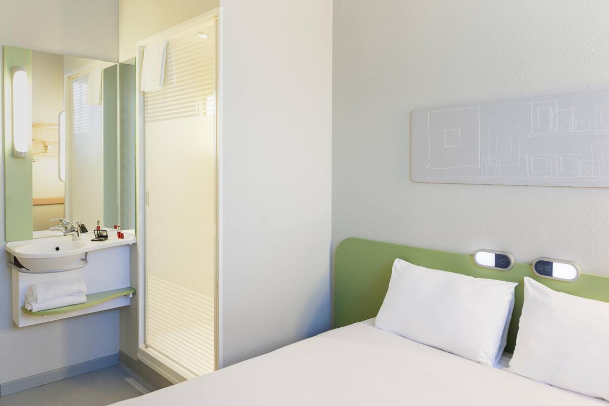 Ein Hotelzimmer mit Bett, Nachttisch, Spiegel, Waschbecken und Handtuch.