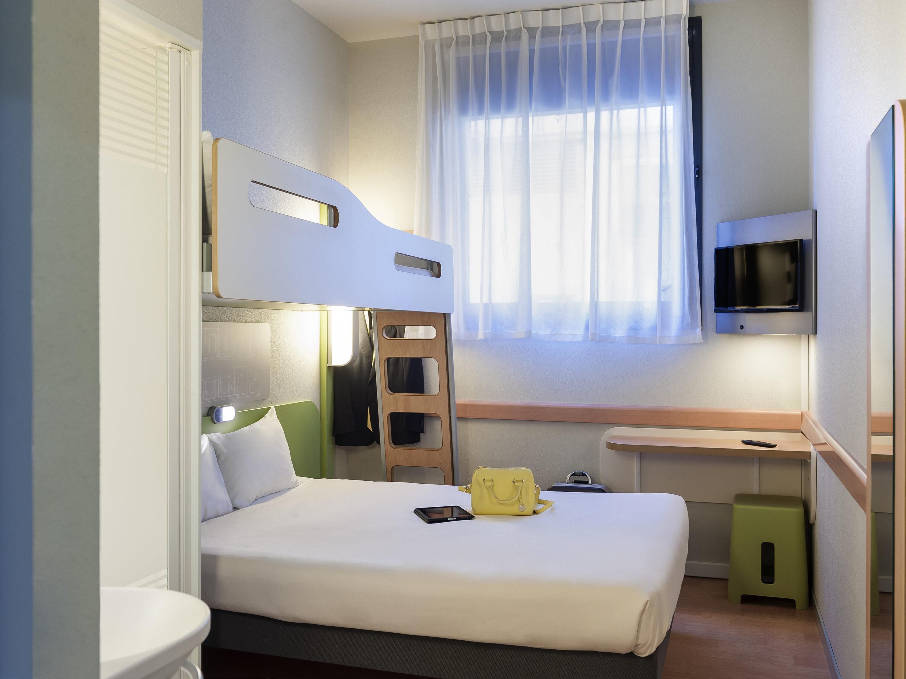 Ein kompaktes Hotelzimmer mit Etagenbett, einem an der Wand montierten Flachbildfernseher, einem kleinen Schreibtisch und einem Fenster mit durchsichtigen Vorhängen.