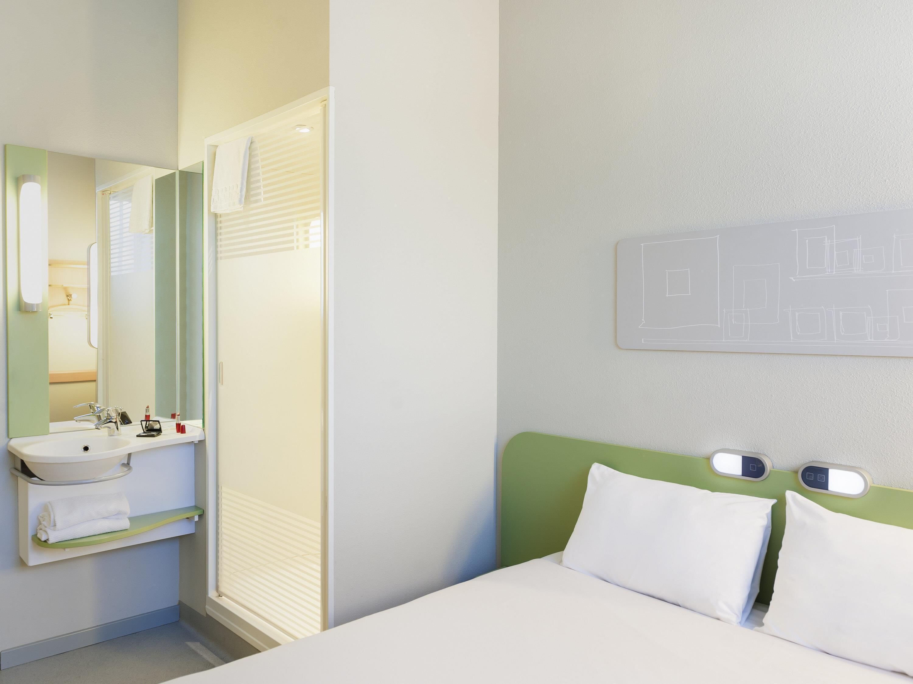 Ein Hotelzimmer mit einem Bett, einem Nachttisch, einem Spiegel, einem Waschbecken und einem Fenster mit Jalousien.