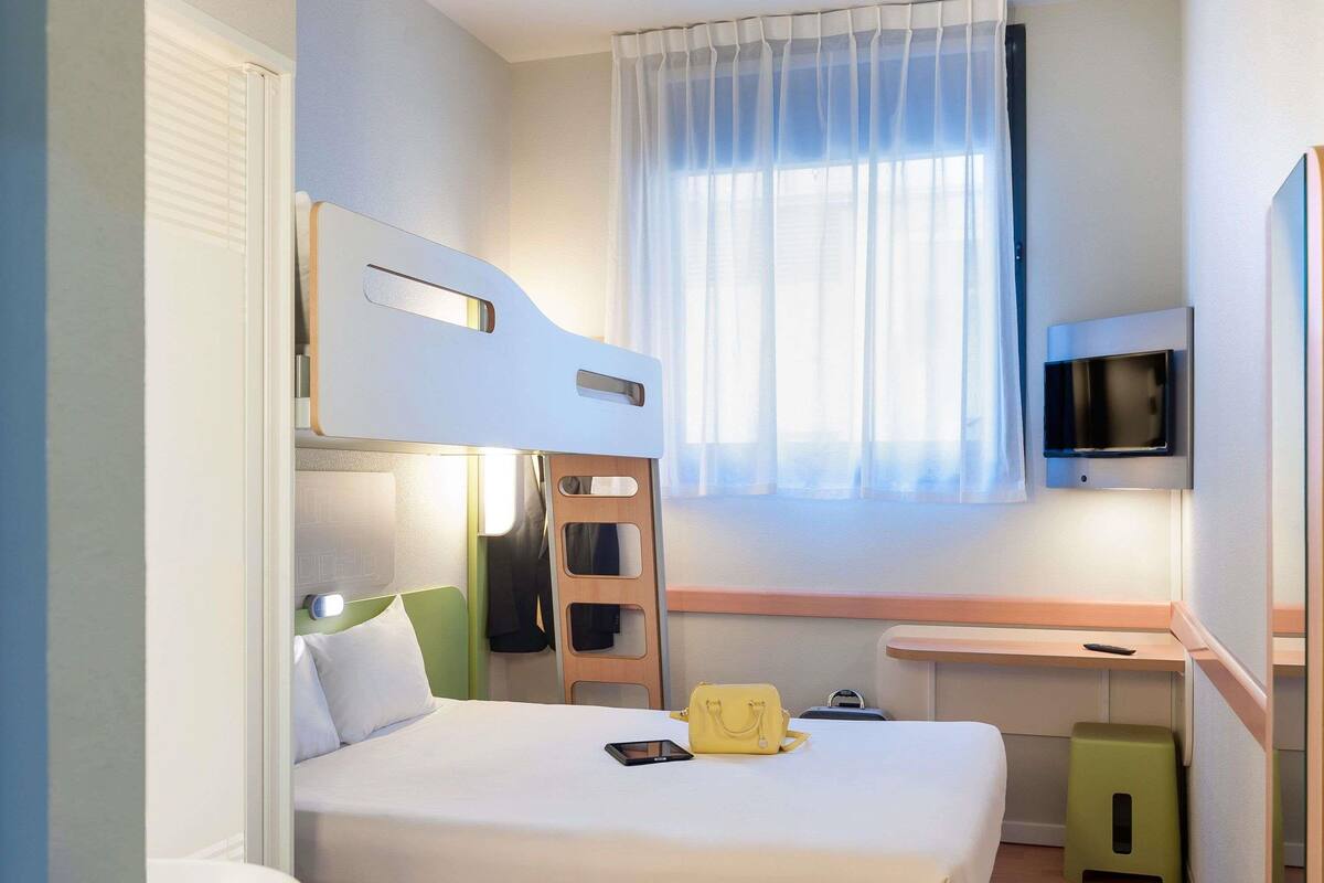 Ein kleines Hotelzimmer mit einem Etagenbett, einem Nachttisch mit Lampe, einem kleinen Hocker, einem an der Wand montierten Fernseher und einem Fenster mit durchsichtigen Vorhängen.