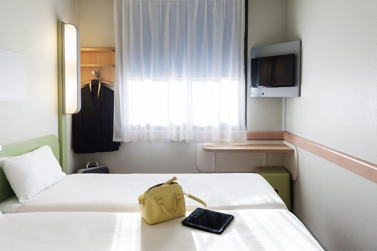 Hotellihuone, jossa on sänky, yöpöytä, televisio, ikkuna verhoilla ja naulakko, jossa on takki.