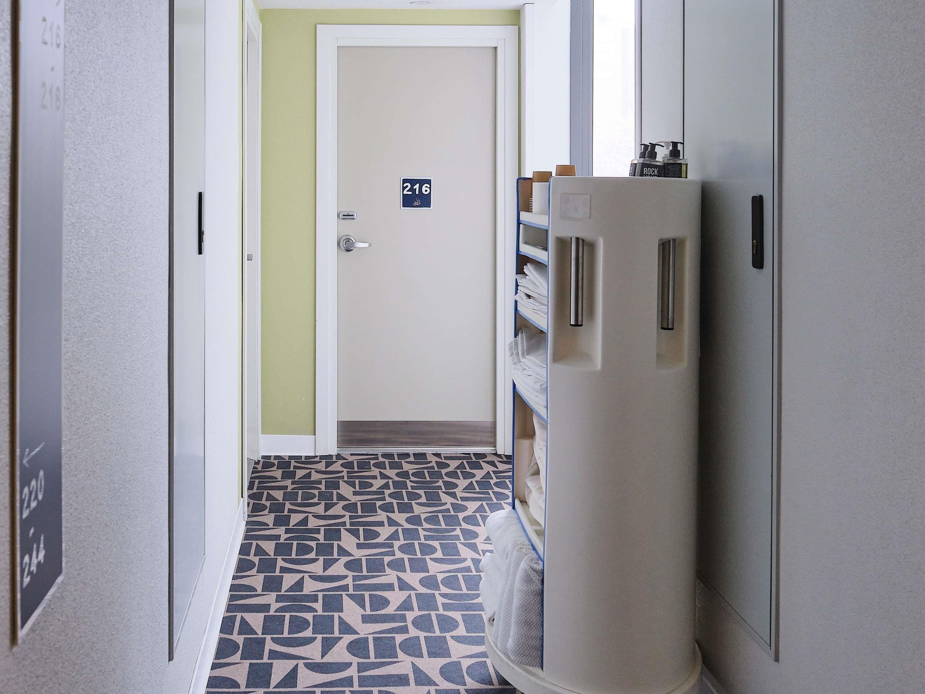 A hallway with a door marked 216, a patterned floor, and a tall cabinet with a bottle on top.