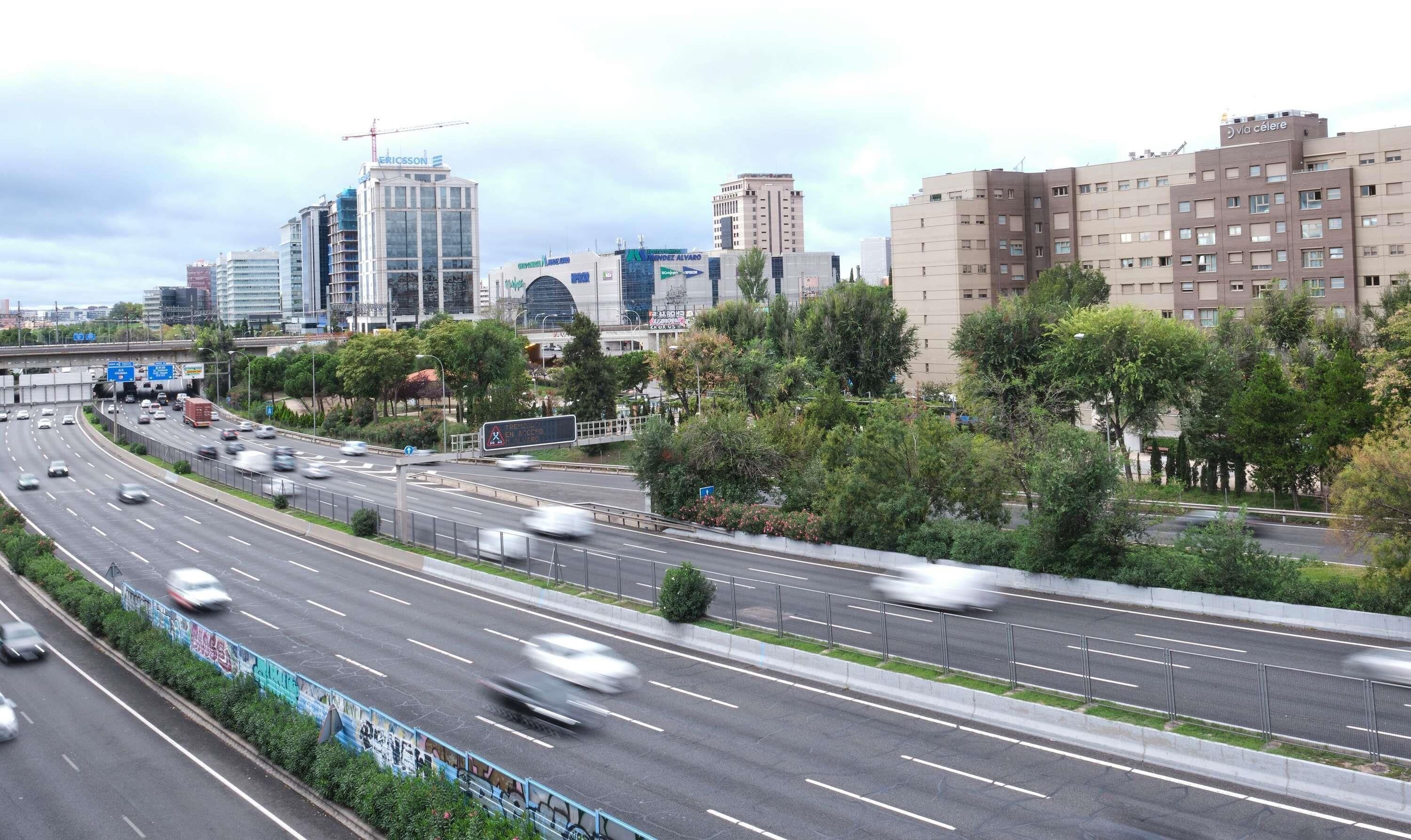 Una autopista con varios carriles y tráfico moderado, rodeada de edificios urbanos y zonas verdes.
