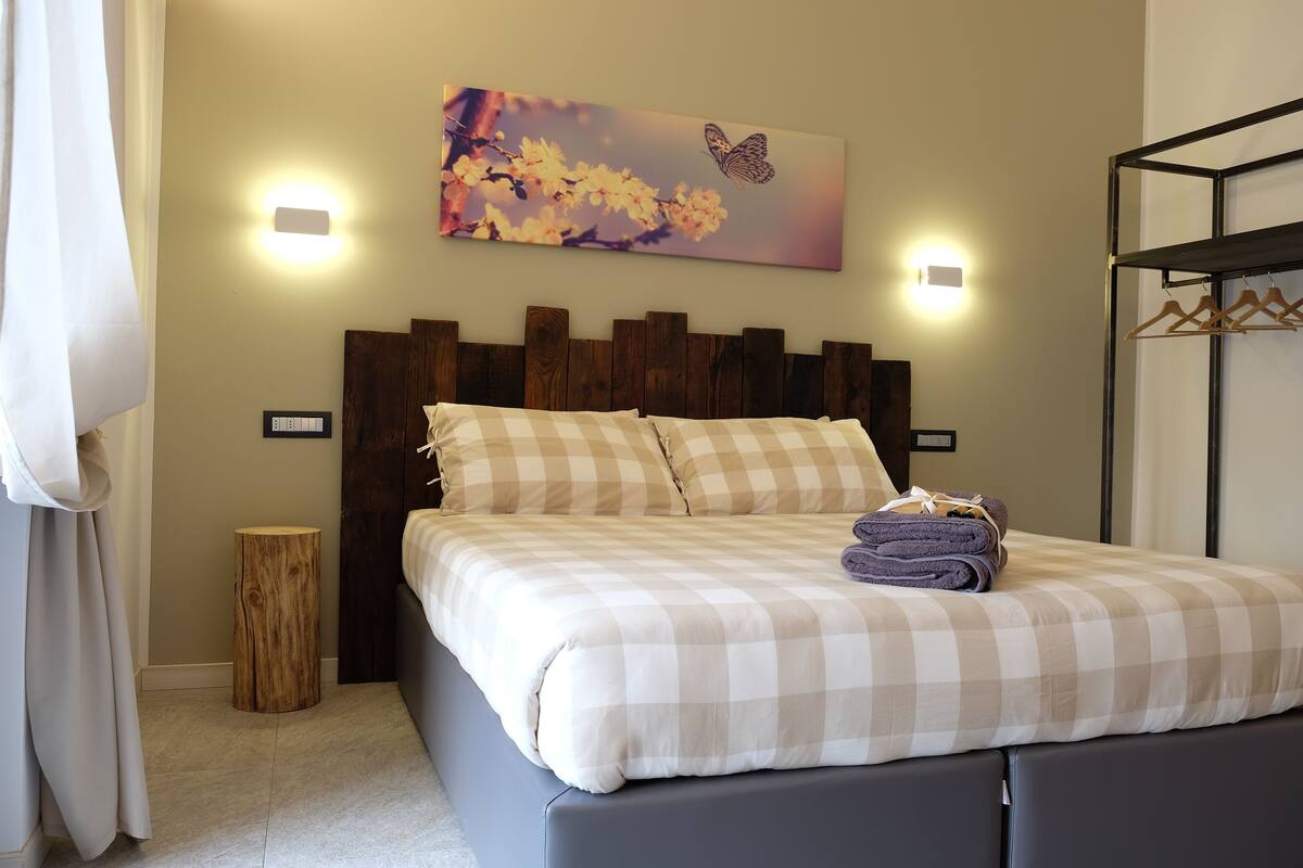 A neatly made bed with a checkered bedspread, a headboard with a wooden panel design, and a wall-mounted painting of flowers and a butterfly.