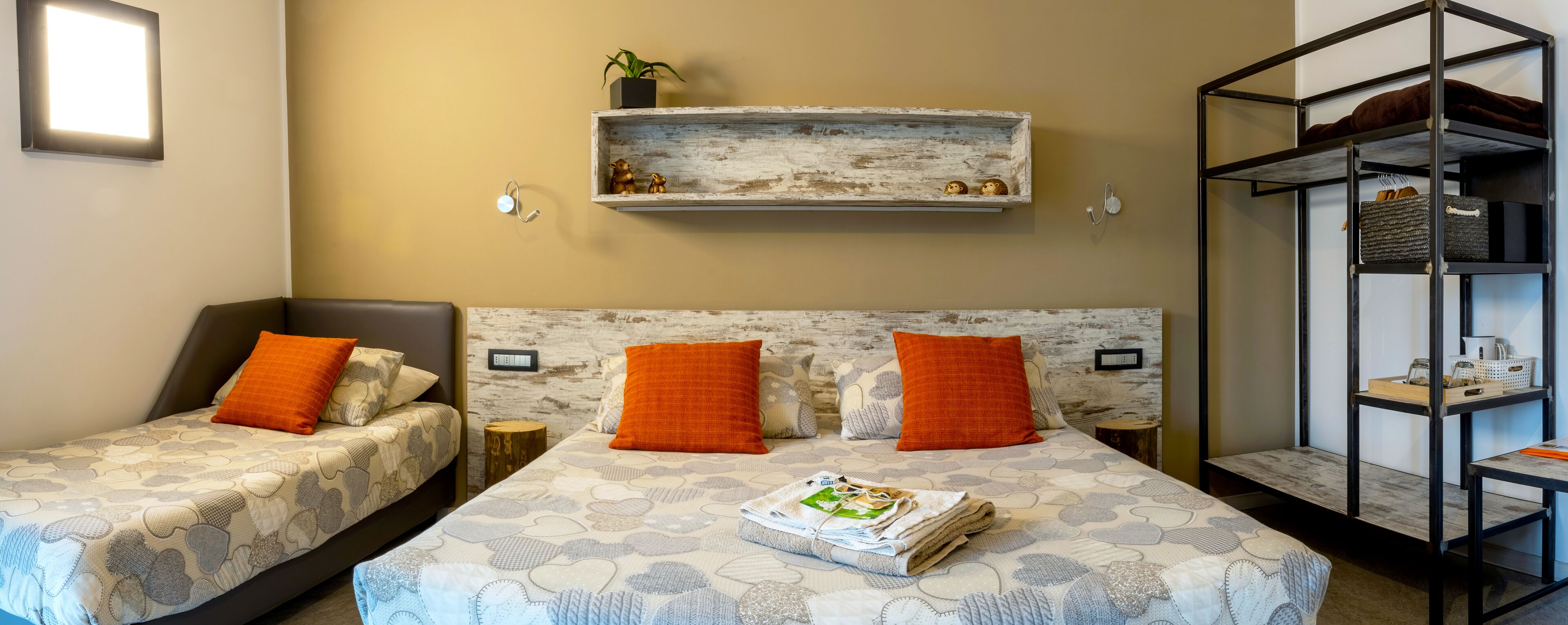 Une chambre d’hôtel avec deux lits, chacun agrémenté de draps à motifs et d’oreillers orange. Une étagère en bois, ornée d’objets décoratifs et d’une petite plante, est placée au-dessus des lits. Un présentoir en métal avec un panier et une bouilloire se trouve sur le côté droit.