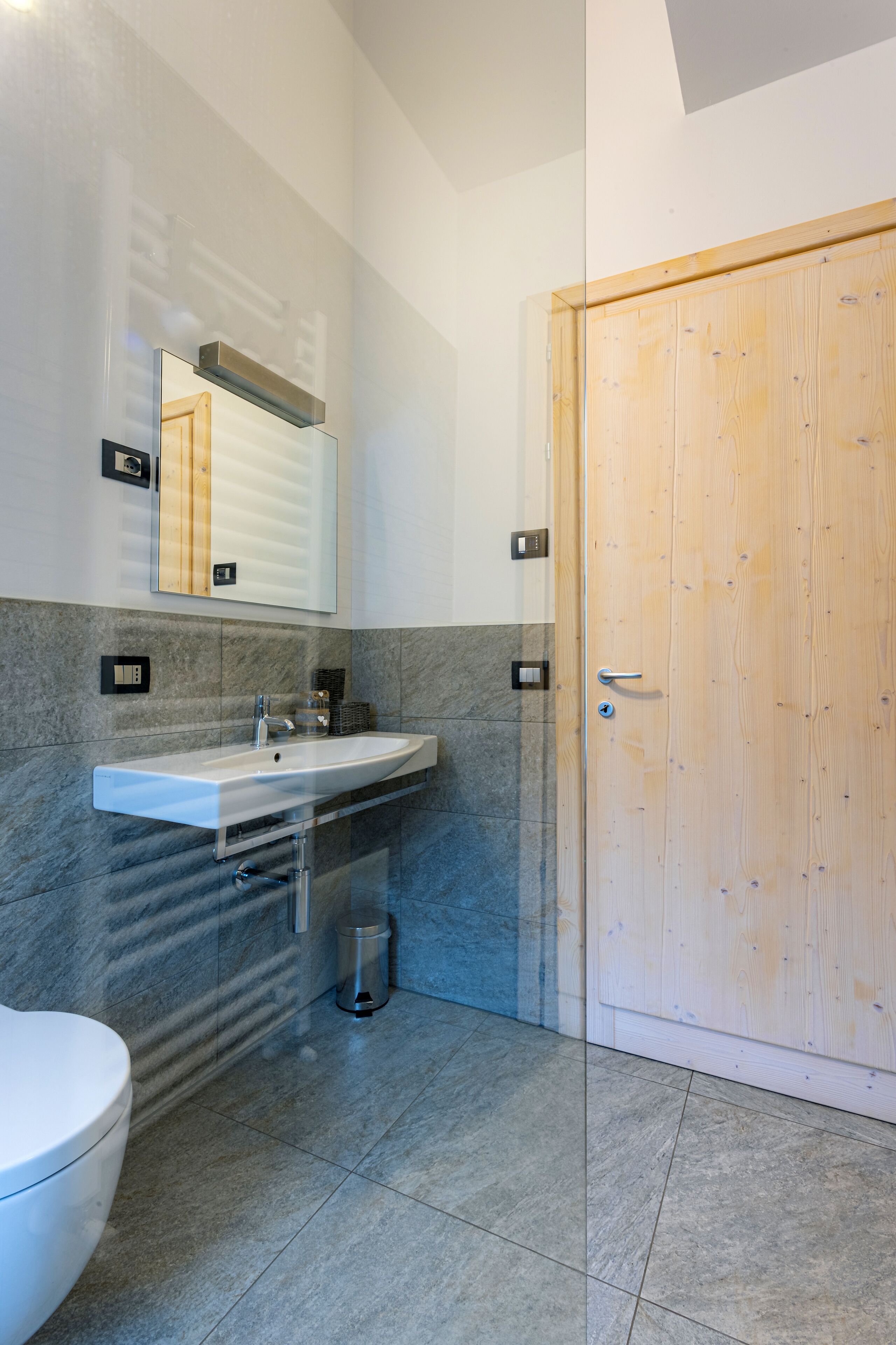 Un bagno moderno con un lavandino bianco, uno specchio e una porta in legno.