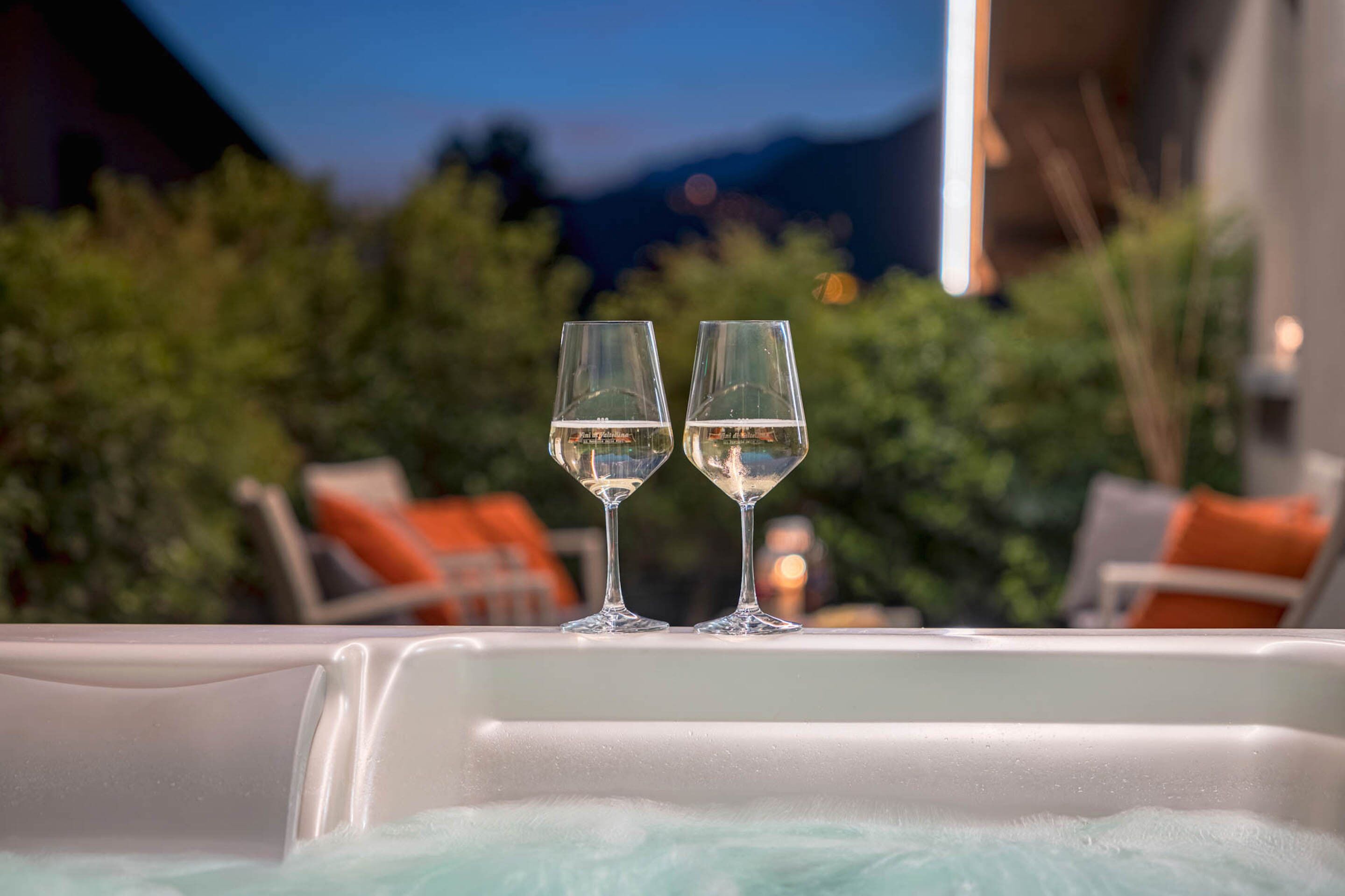 Two glasses of white wine on a white table with a view of greenery and mountains in the background.