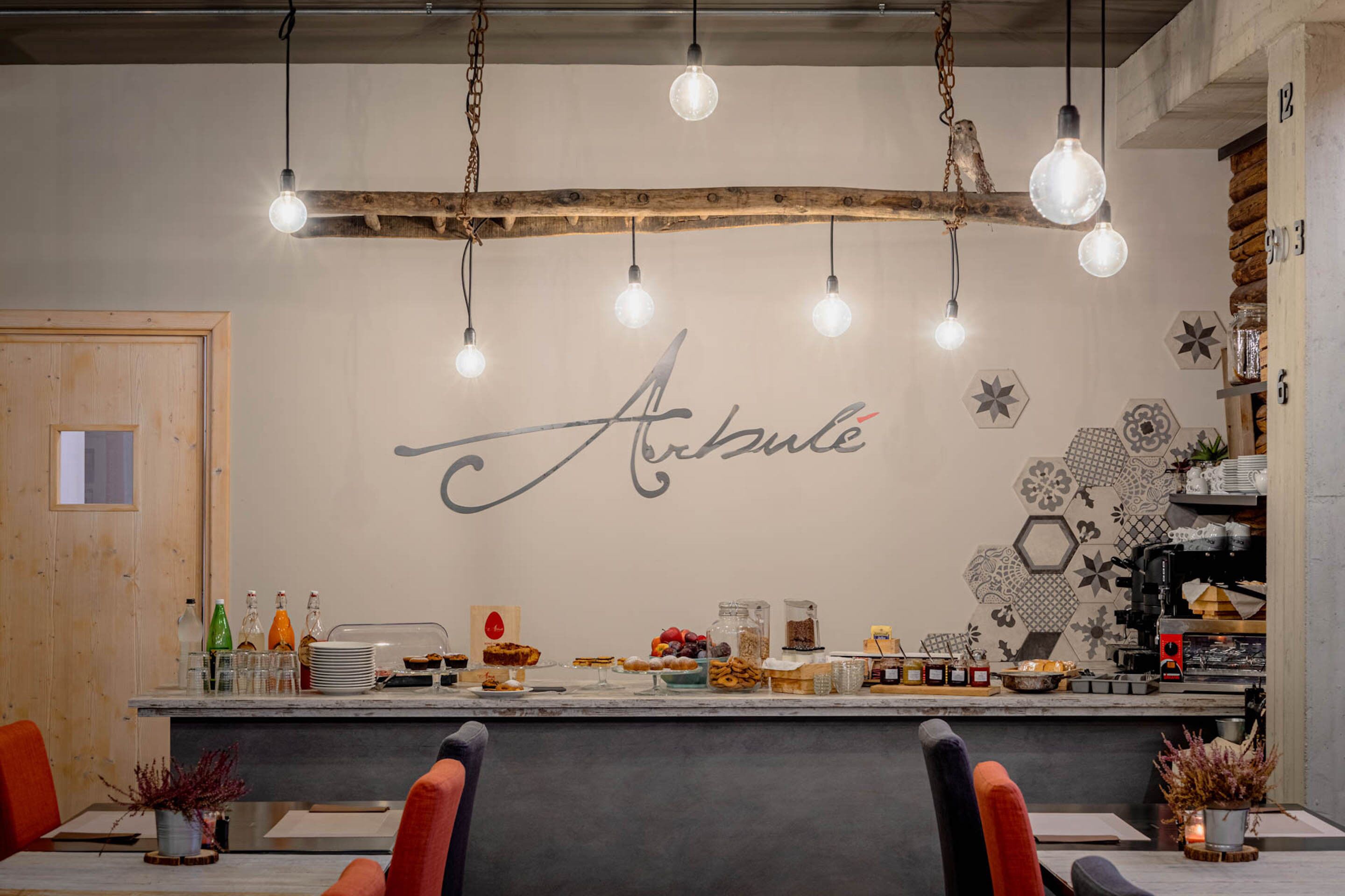 Interior de restaurante con mostrador de bar de madera, bombillas colgantes y una pared con un gran letrero con escritura cursiva.