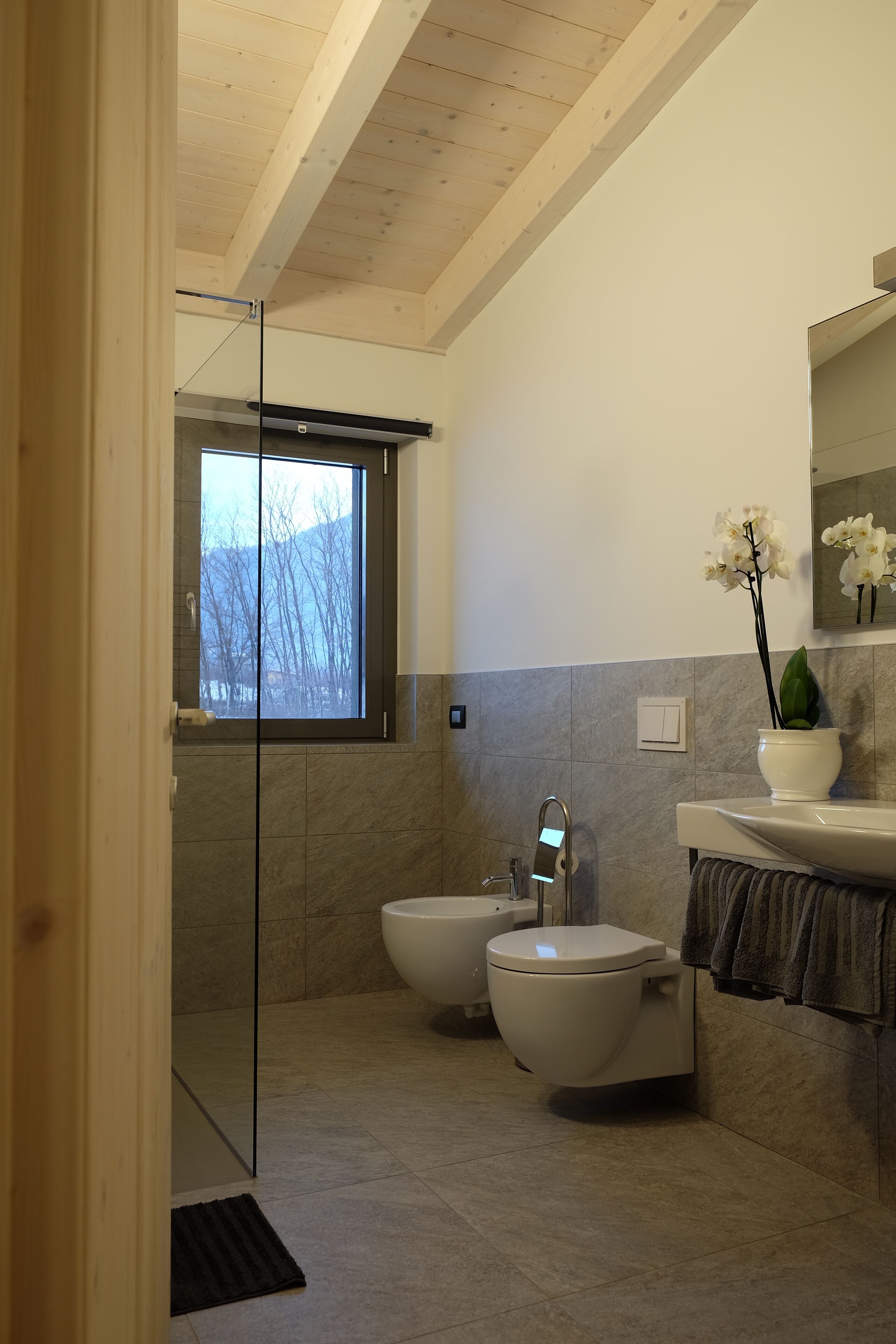 Un baño moderno con cabina de ducha de vidrio, un bidé, inodoro, lavamanos y ventana que proporciona luz natural.