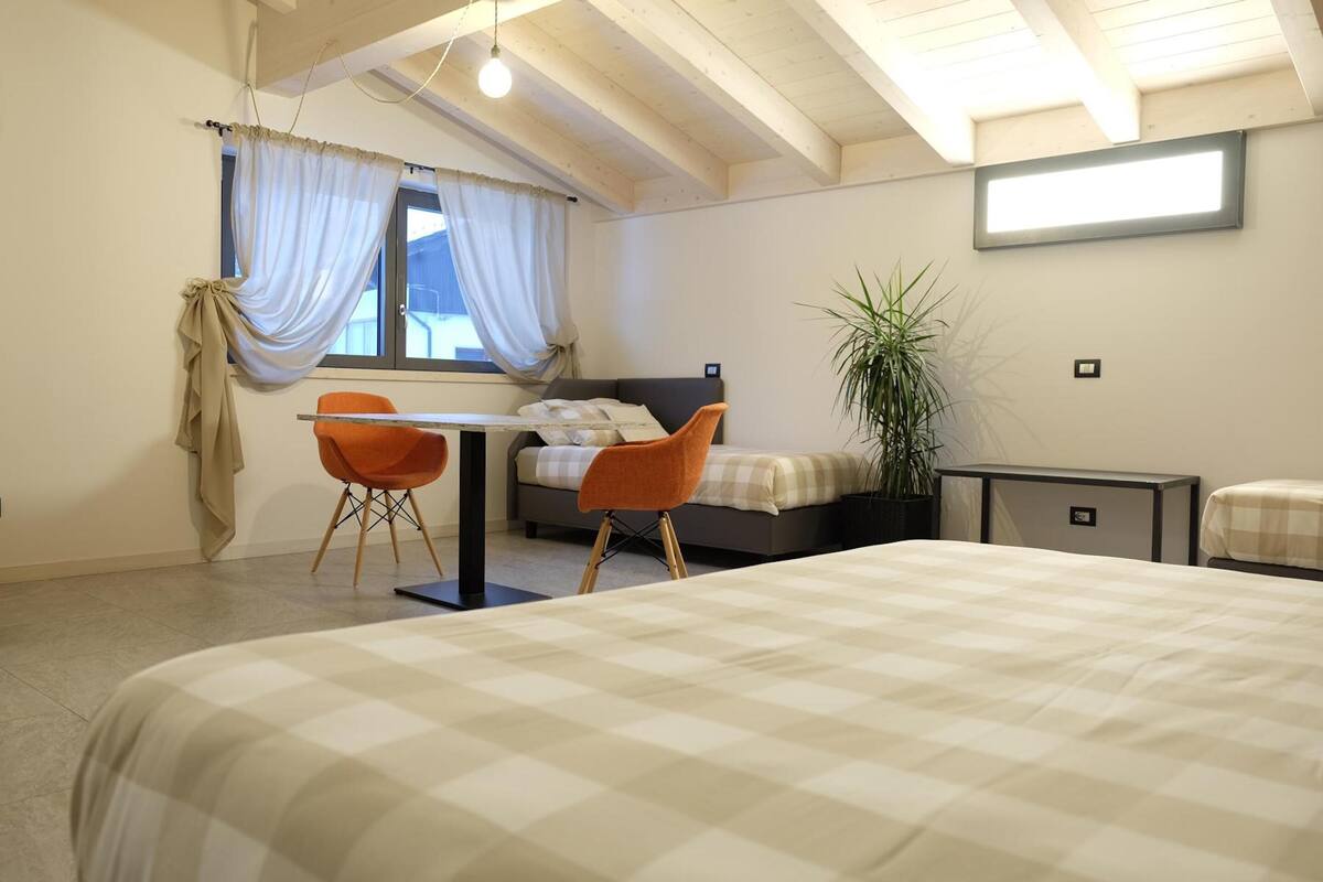 A bedroom with a bed, a desk, two chairs, a bedside table, a potted plant, and a window with curtains.