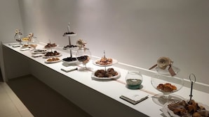 Una larga mesa buffet blanca con diferentes alimentos expuestos en recipientes de vidrio y platos, frente a una pared lisa.