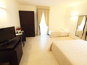 A hotel room with a bed, a desk with a TV, a chair, a small table with a vase of flowers, and a window with curtains.